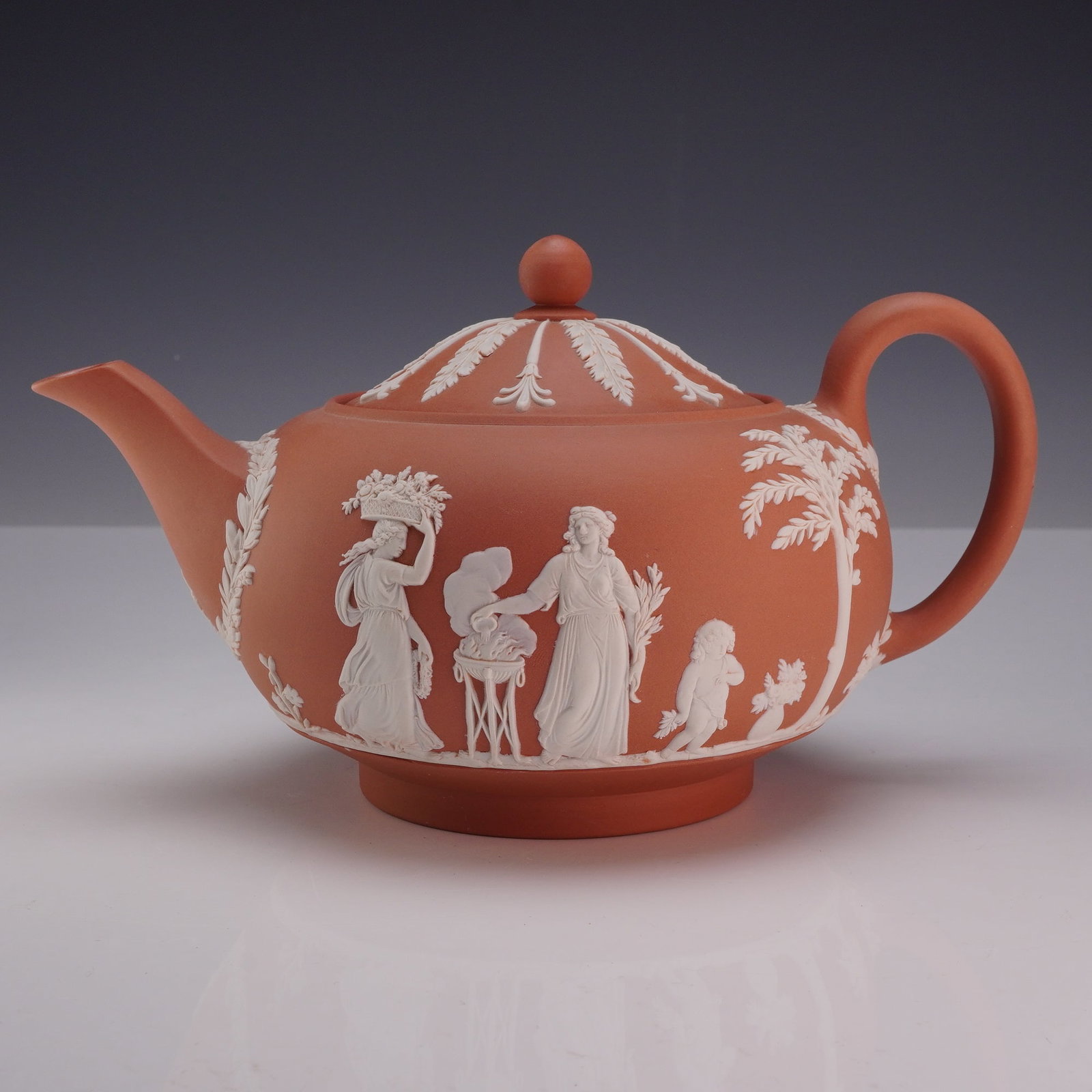Wedgwood Rosso Antico Jasperware Lidded Teapot with Classical Relief Decoration: This Wedgwood Jasperware lidded teapot is crafted from Rosso Antico stoneware, a dry-bodied, unglazed terracotta-colored ceramic. The bulbous body features white sprigged bas-relief classical motifs d