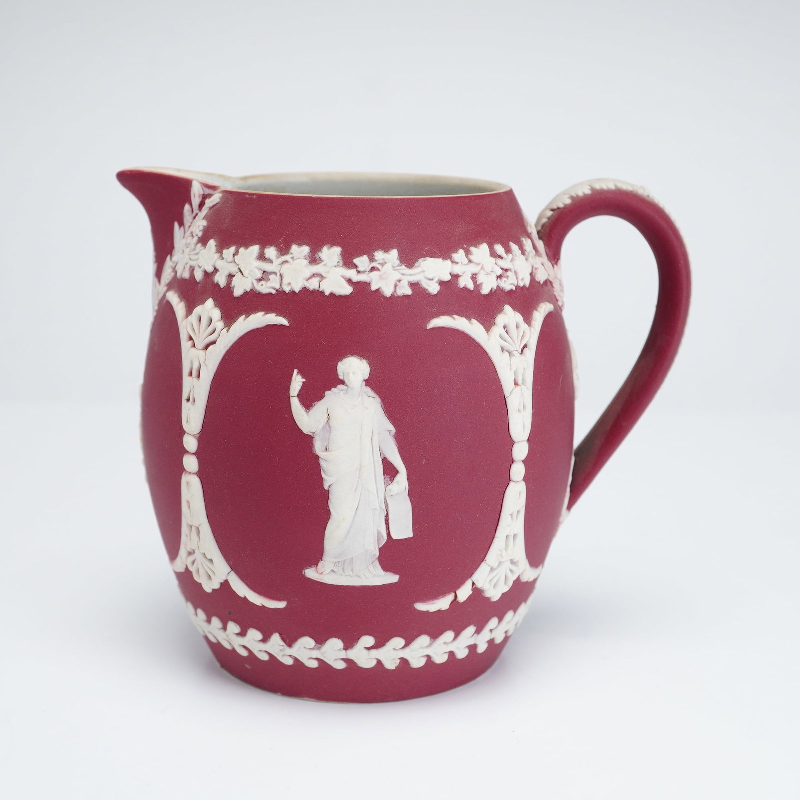 Wedgwood Crimson Jasperware Cream Jug: This Wedgwood crimson dip jasperware cream jug features a matte-finish stoneware body with applied white bas-relief decoration. The vessel is designed with an ovoid body, a pinched spout, and a loop h