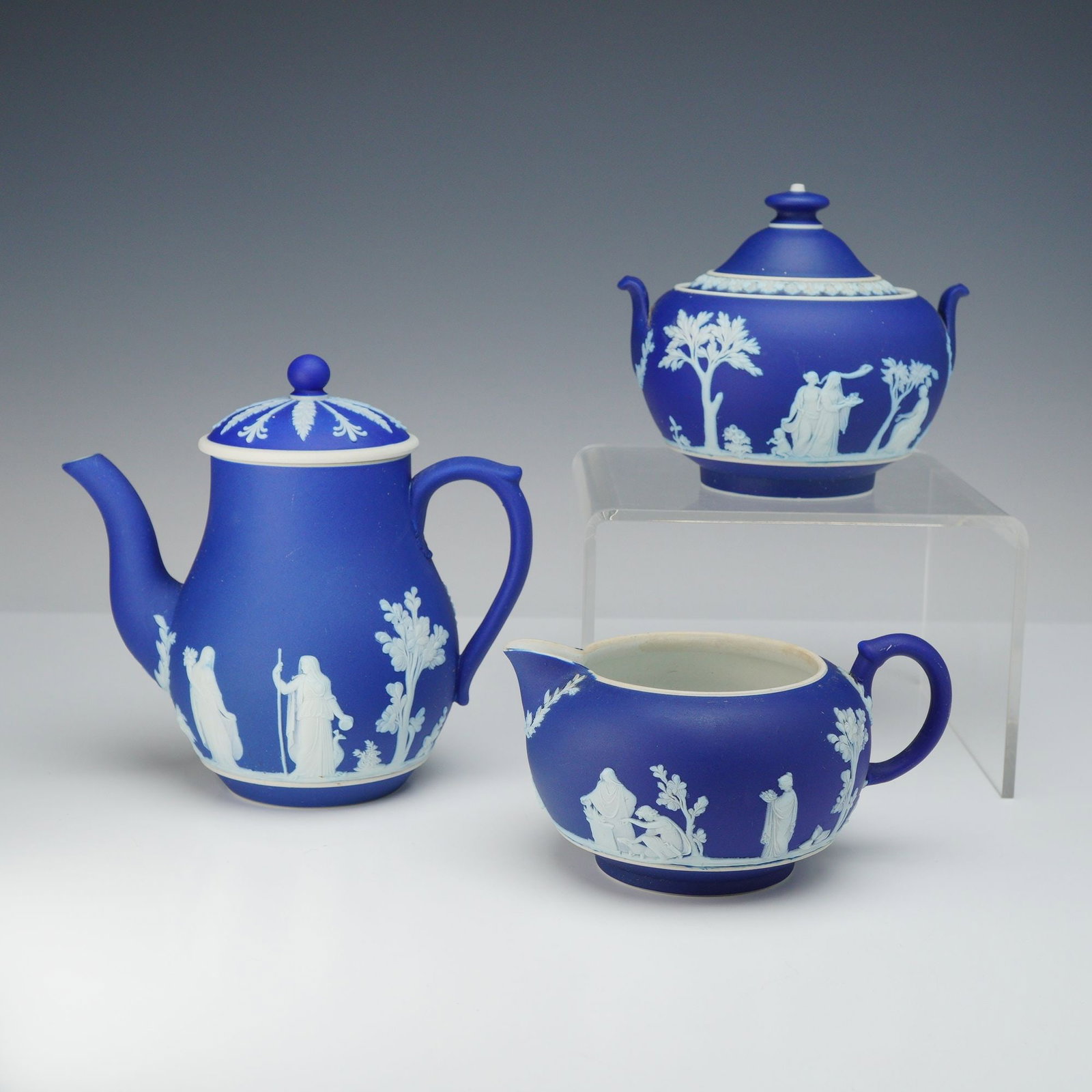 3pc Wedgwood Dark Blue Jasperware Coffee Pot, Creamer, and Sugar Bowl: A three-piece Wedgwood dark blue jasperware service set featuring traditional Neoclassical white sprigged bas-relief decorations. The set consists of a tall, slender lidded coffee pot with a steam ven