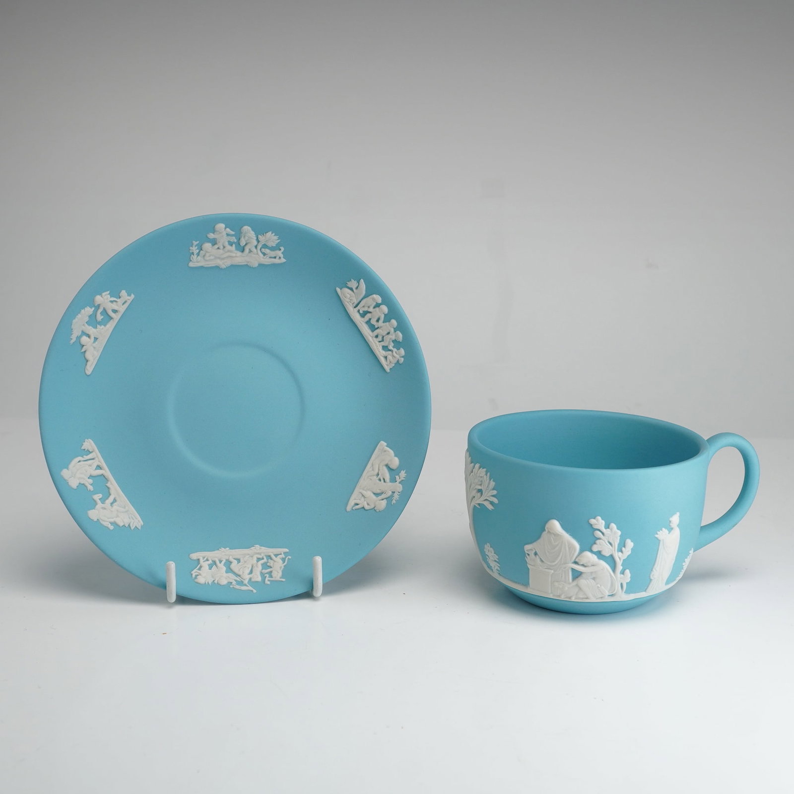 2pc Wedgwood Light Blue Jasperware Teacup and Saucer: This set consists of a Wedgwood light blue jasperware teacup and saucer featuring sprigged white bas-relief neoclassical decorations. The teacup displays various figures in draped garments alongside s