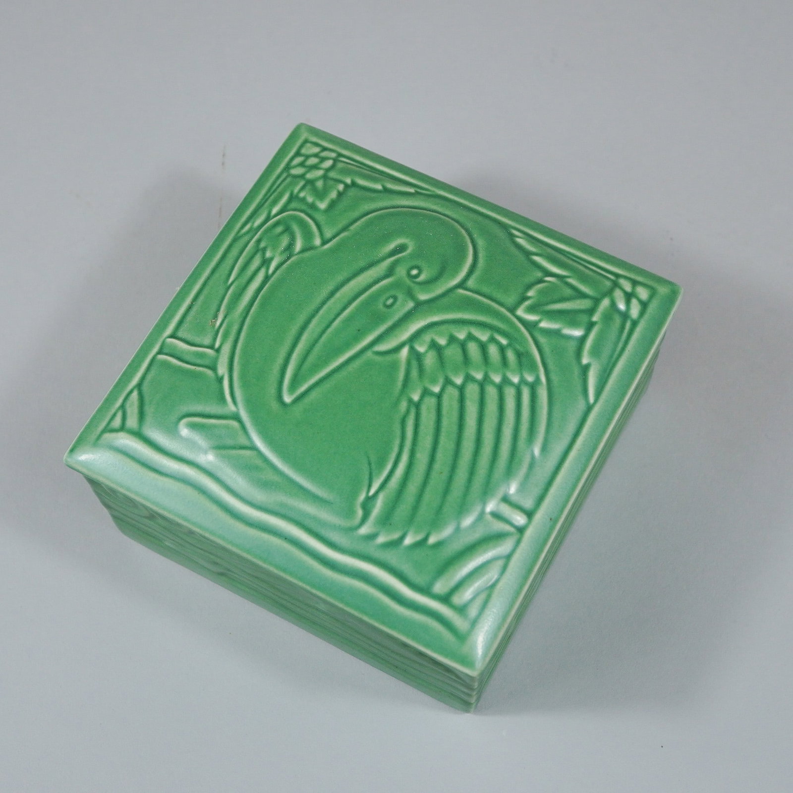 Wedgwood Green Glazed Earthenware Pelican Square Box with Lid: This square lidded box is a piece of Wedgwood glazed earthenware, finished in a monochromatic green glaze. The lid features a molded relief design of a stylized pelican positioned amidst foliage and s