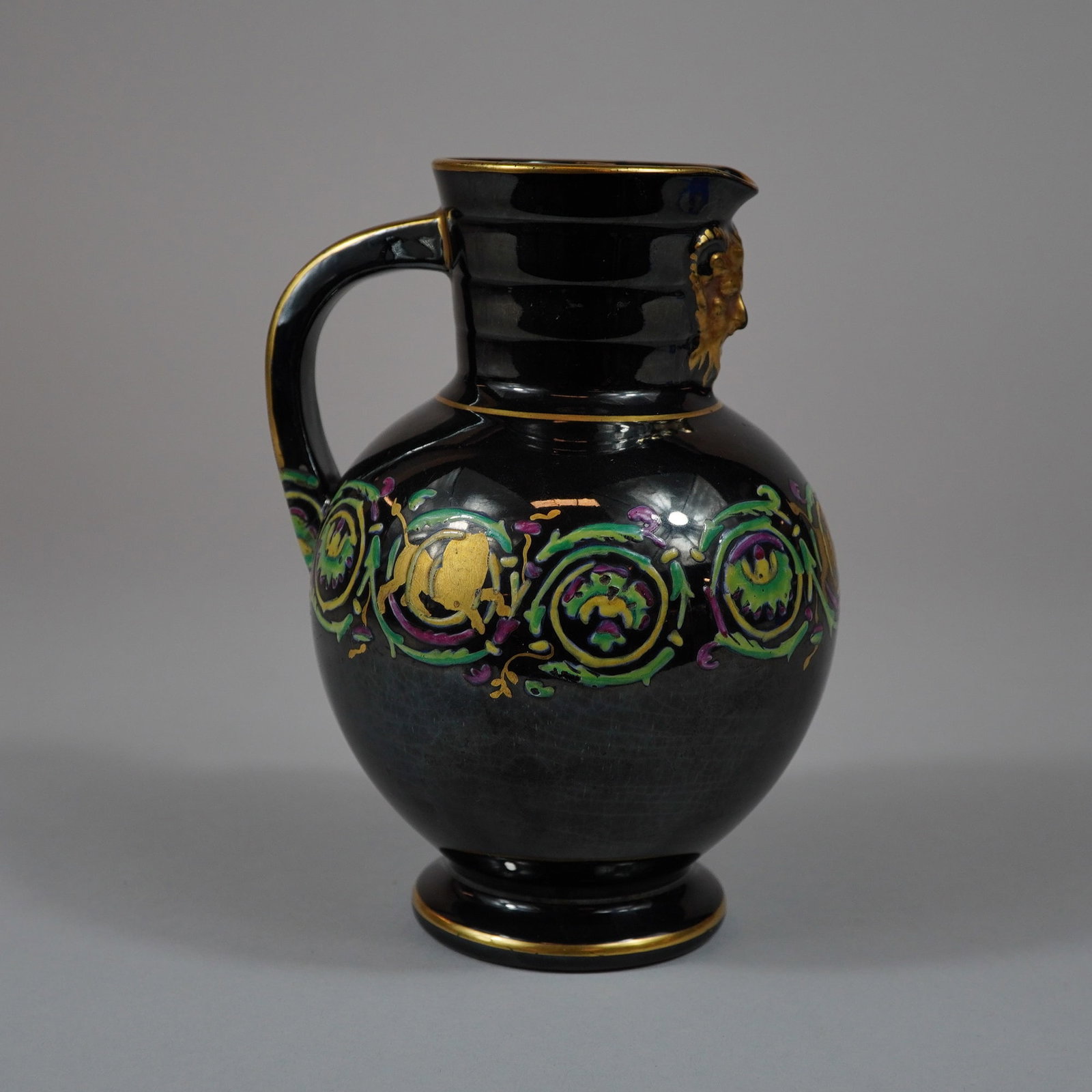 Black Glazed Earthenware Pitcher with Gilded Satyr Mask and Lion Rampant: This glazed earthenware pitcher is designed in the Renaissance Revival style. The vessel features a bulbous baluster-form body supported by a short, flared foot. Its cylindrical neck is detailed with 