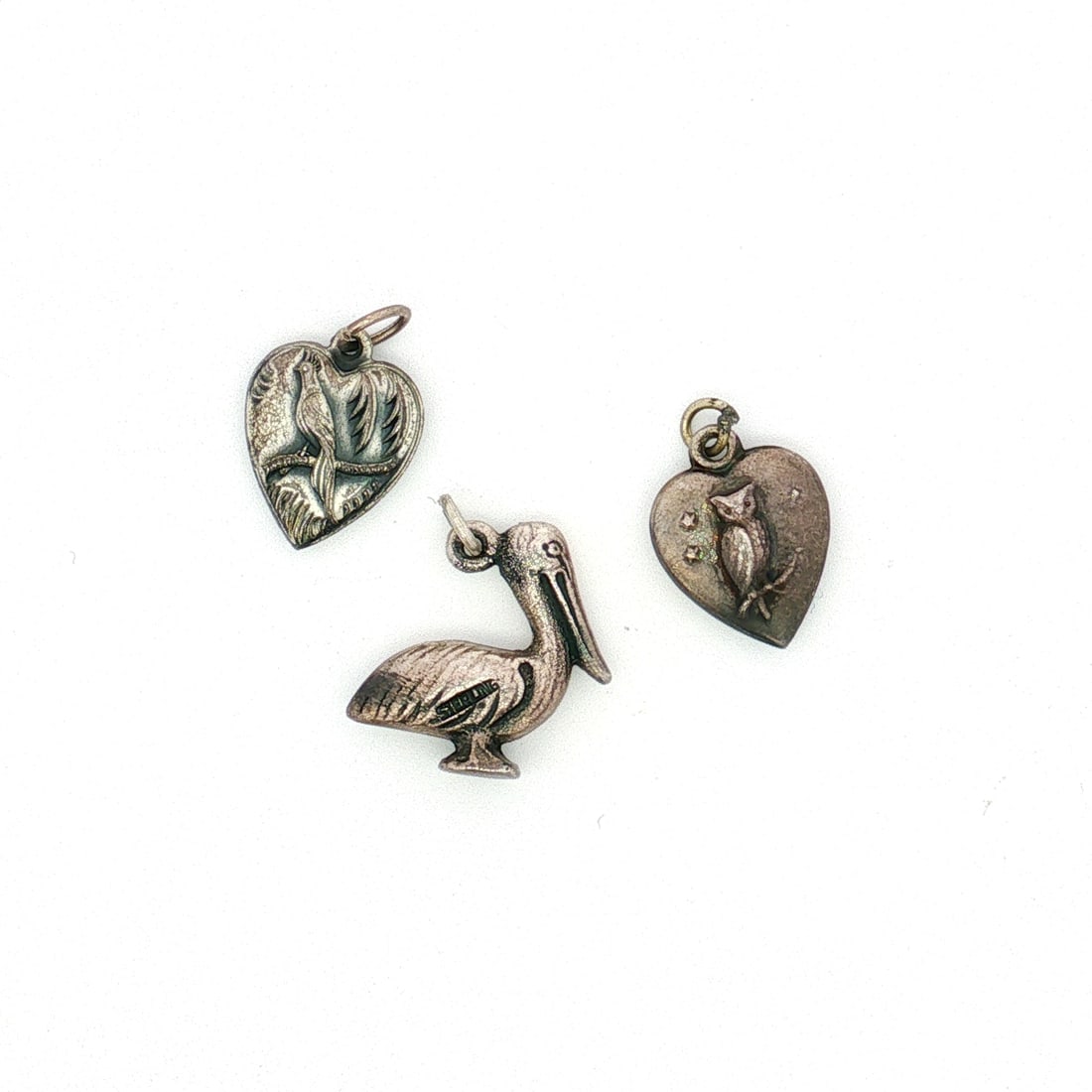 3pc Sterling Silver Bird Charms: A group of sterling silver charms with bird-themed designs. The lot includes two heart-shaped charms, one featuring an owl and the other a bird with long tail feathers. Also included is a pelican char