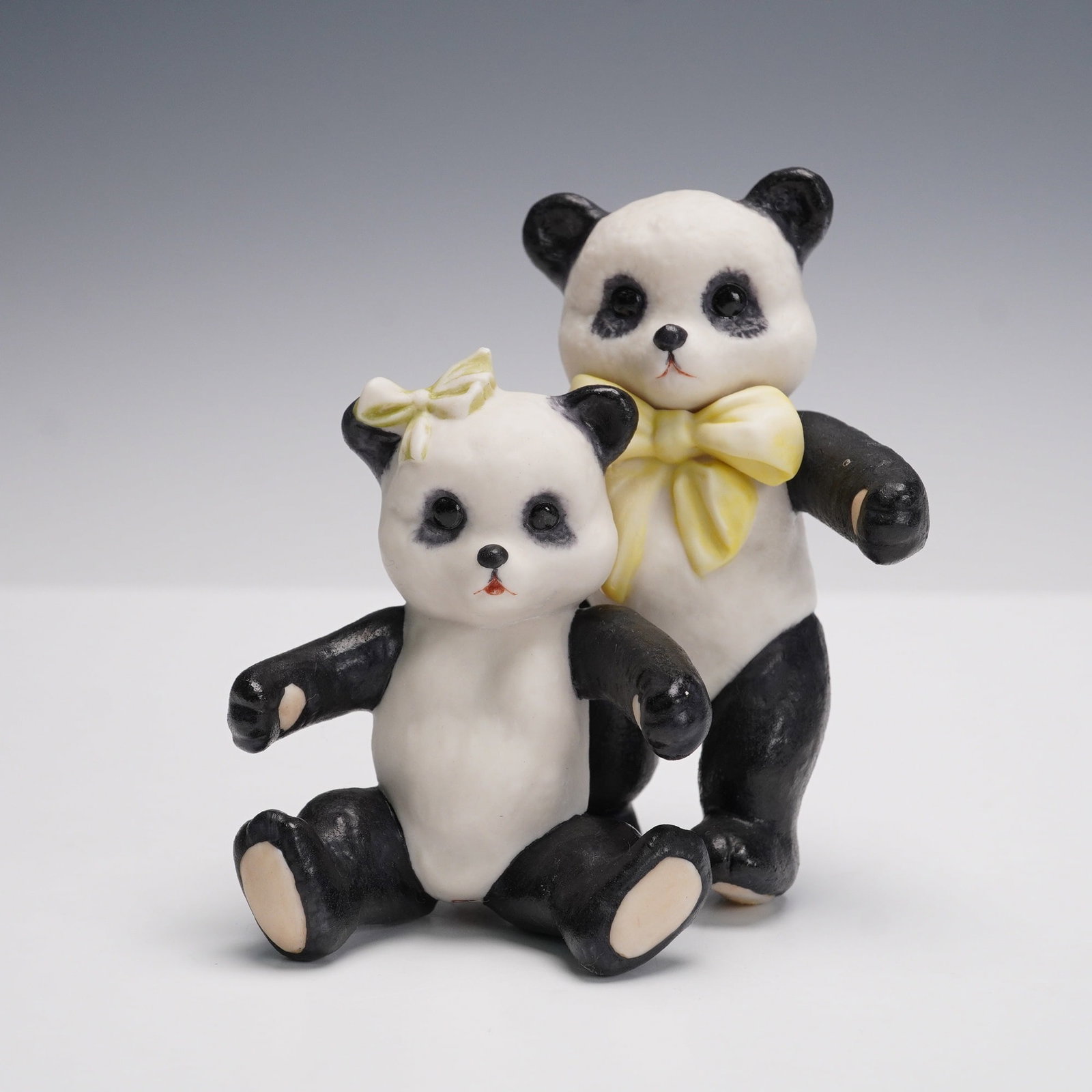 Cybis Porcelain Panda Pair with Bows Figurine: Delightful Cybis porcelain figurine featuring two pandas posed together, each adorned with a bow - yellow at the neck and green at the ear. The hand-painted details enhance their playful expressions a