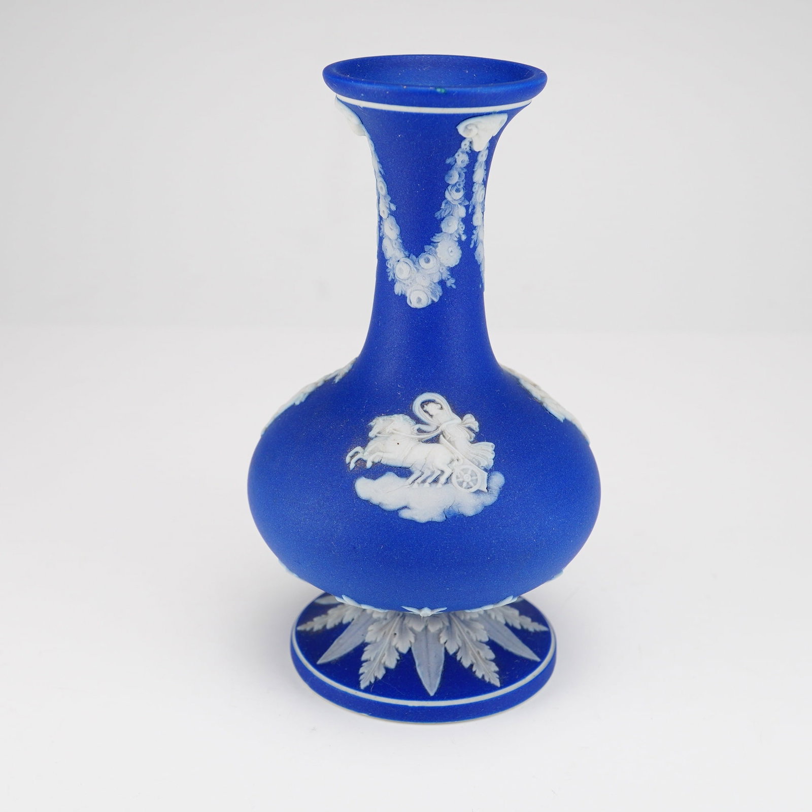 Wedgwood Cobalt Blue Jasperware Footed Vase with Classical Relief: A Wedgwood cobalt blue jasperware footed vase featuring white sprigged neoclassical relief decoration. The vessel is constructed from fine-grained unglazed stoneware with a bulbous body, long tapering