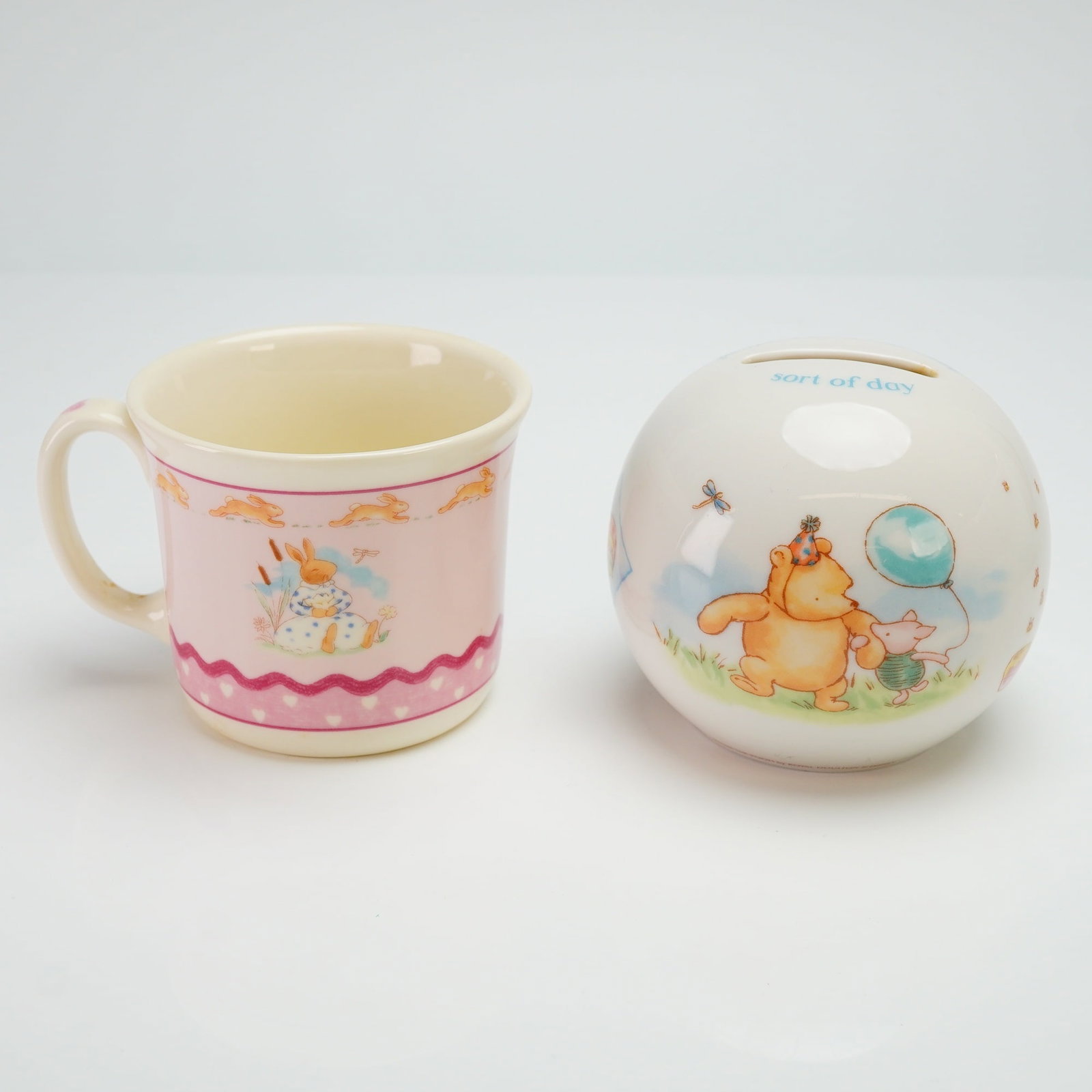 2pc Royal Doulton Bunnykins Mug and Winnie the Pooh Coin Bank: The first item is a Royal Doulton Bunnykins mug in the Sweet Hearts pattern, crafted from bone china. The exterior features a pink ground decorated with illustrations of anthropomorphic rabbits and a 