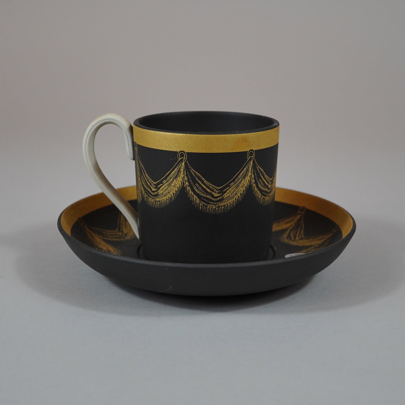 Wedgwood Black Basalt Gilded Teacup and Saucer Set: A matching pair comprising a Wedgwood black basalt stoneware coffee cup and saucer with neoclassical gilded ornamentation. Both pieces are made from unglazed, matte black basalt stoneware. The cup is 