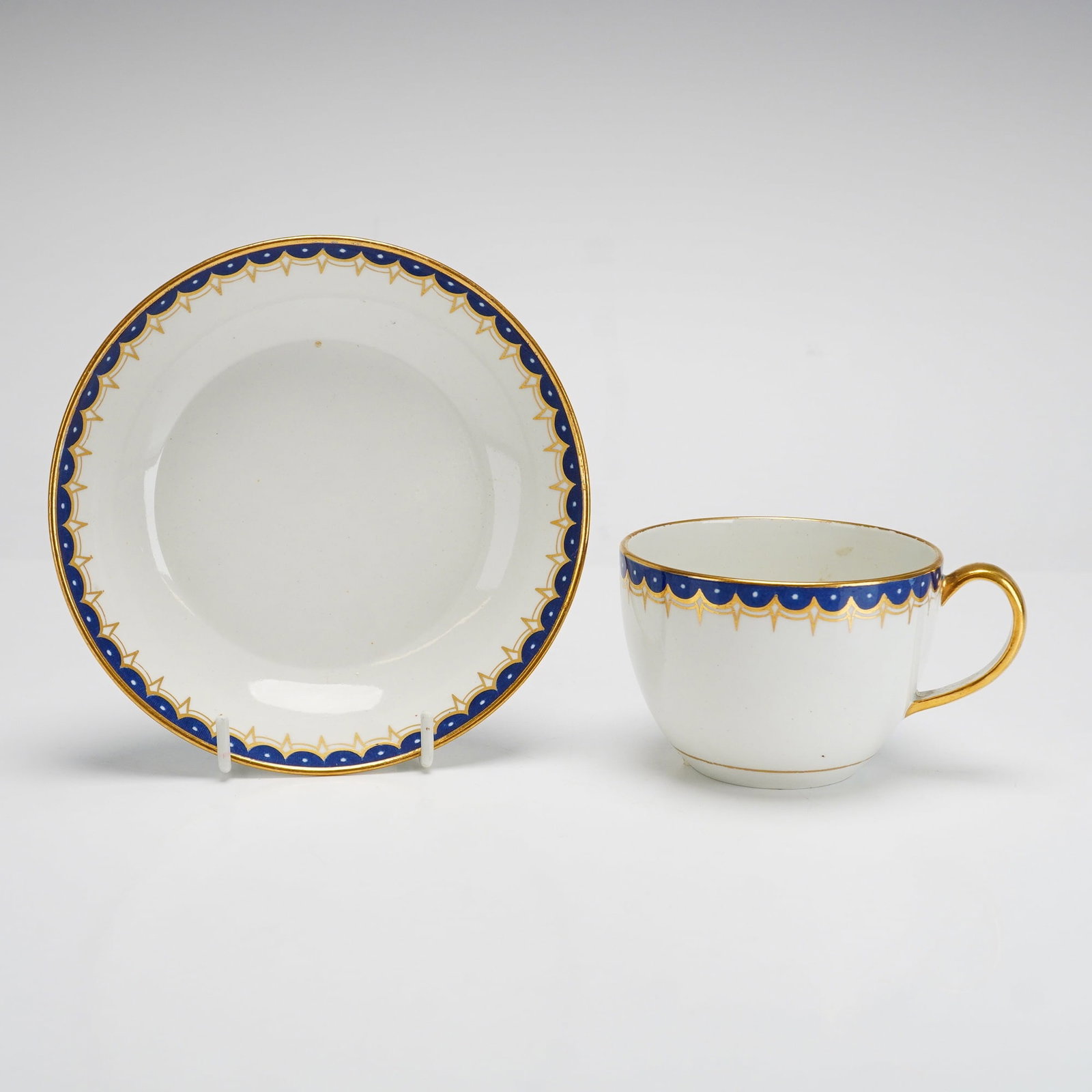 Wedgwood Porcelain Teacup and Saucer with Gilded Cobalt Scalloped Border: This set consists of a fine porcelain teacup and matching saucer manufactured by Wedgwood. The decoration features a refined neoclassical border with a cobalt blue band and a repeating scalloped edge.