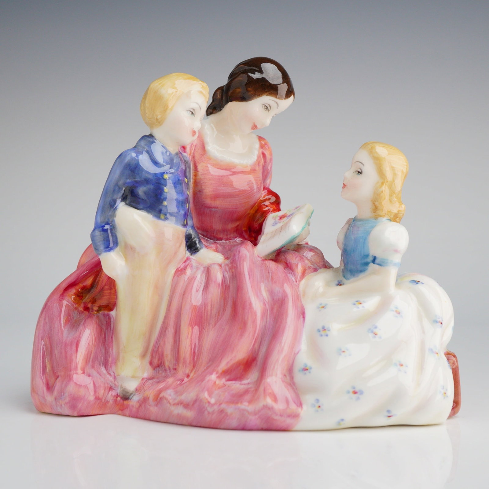 Royal Doulton Figurine, Bedtime Story HN2059: The piece depicts an adult woman seated, reading from a small book or sheet music to a young boy and a young girl seated beside her. The figures are highly glazed and hand-painted, featuring brightly 