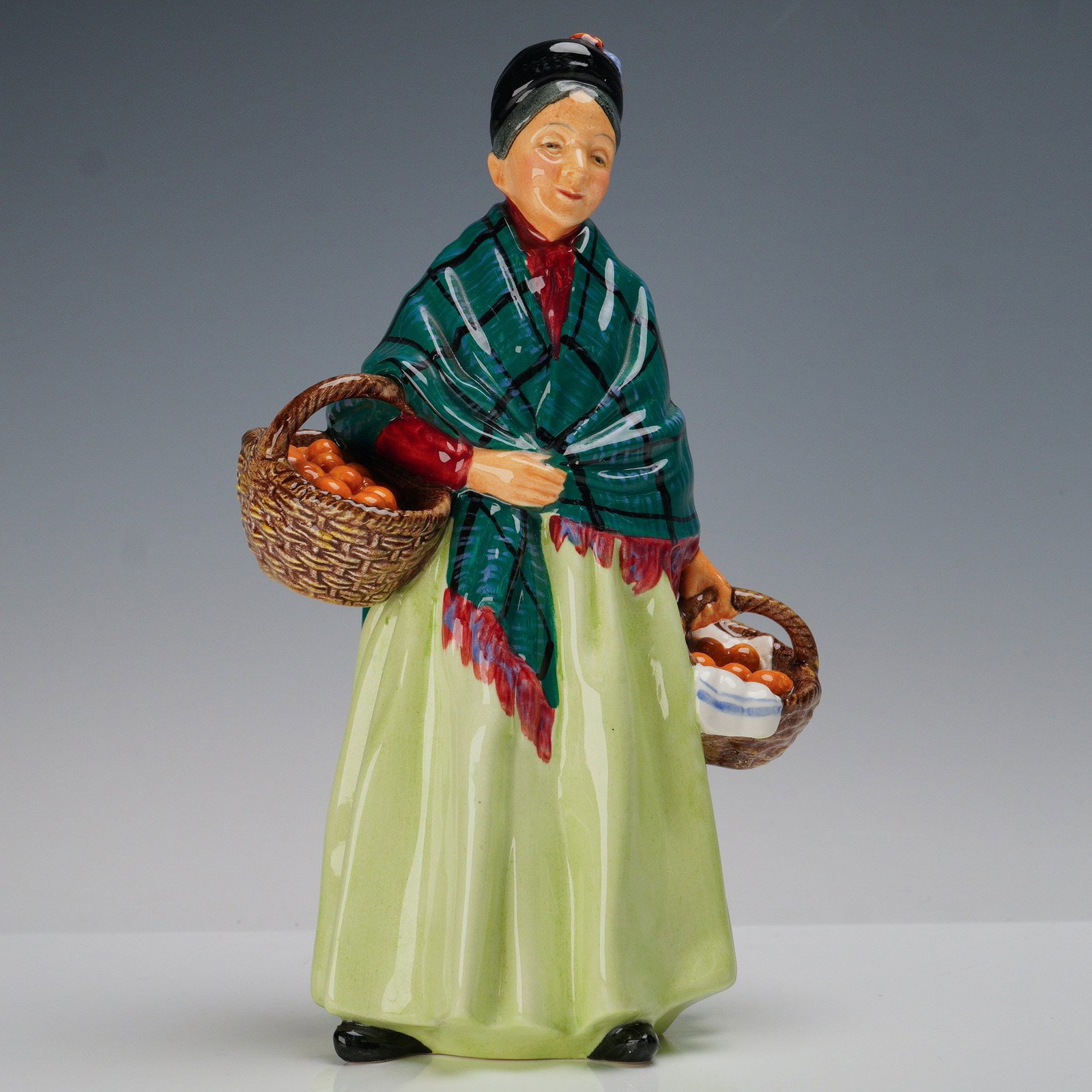 Royal Doulton Figurine, Orange Lady HN1953: This hand-painted porcelain figure depicts a woman carrying baskets filled with oranges. She wears a long pale green skirt, red blouse, and a green checked shawl with red fringe, while her dark hair i