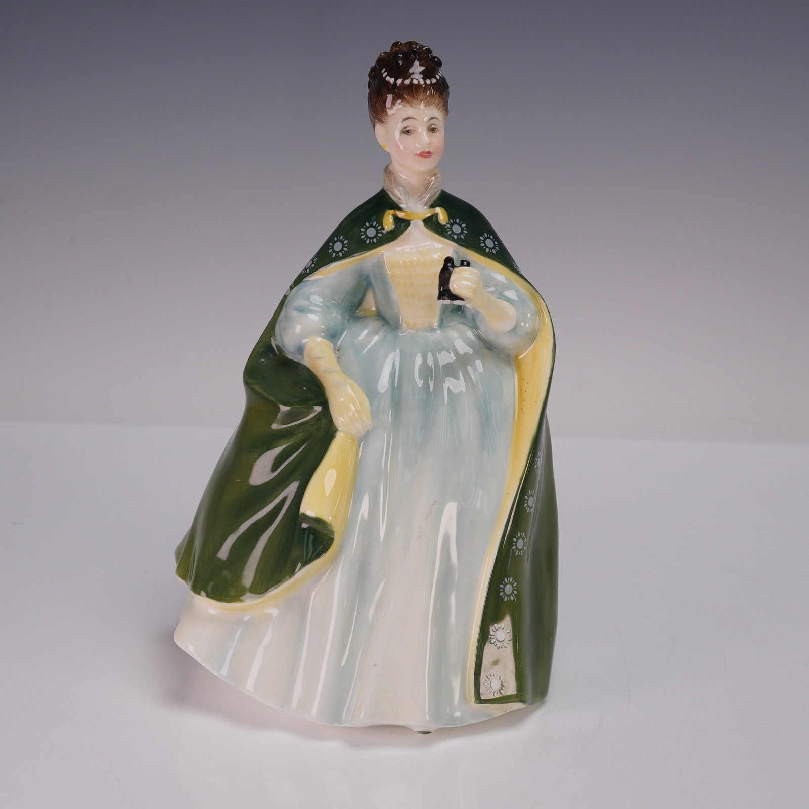 Royal Doulton Figurine, Premiere HN2343: A finely detailed Royal Doulton figurine titled Premiere, model HN2343, designed in 1969. The piece depicts a finely dressed lady in a flowing pale gown with a deep green cloak lined in yellow, holdin