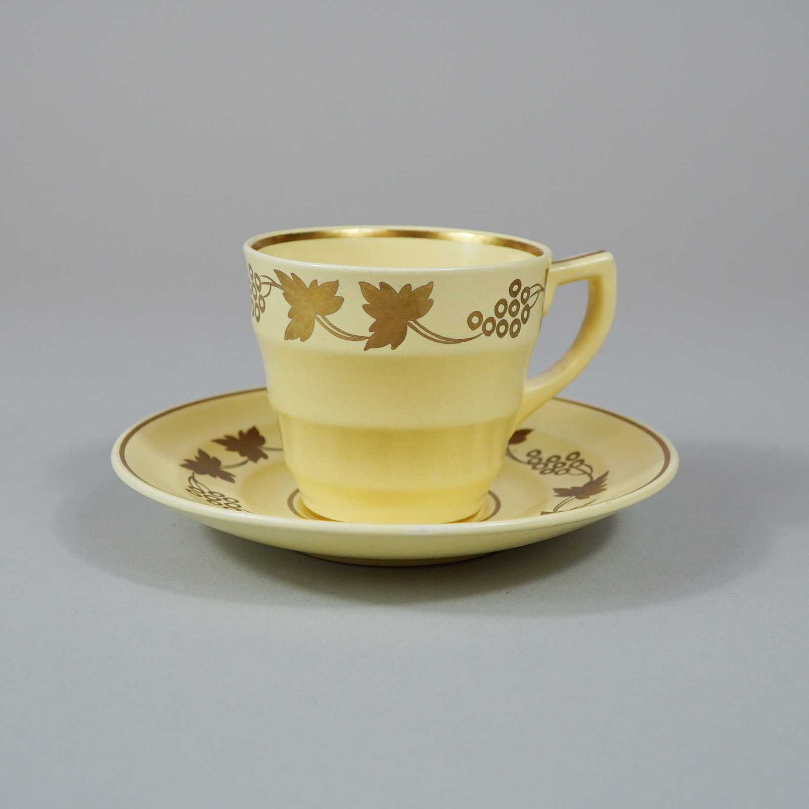 2pc Wedgwood Matt Straw Glazed Cup and Saucer with Gilt Grapevine Pattern: This cup and saucer set was produced by Wedgwood at the Etruria works in England. The set is made of earthenware and features a Matt Straw pale yellow matte glaze decorated with a gold luster grapevin