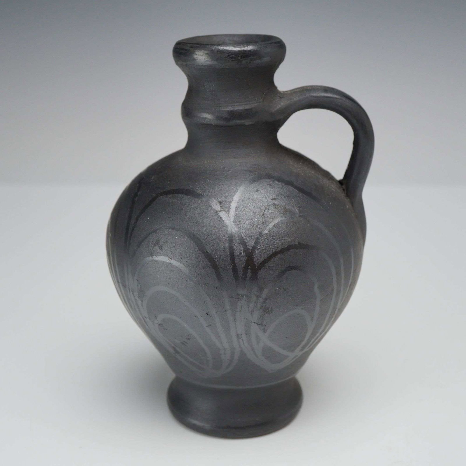 Romanian Burnished Black Earthenware Pitcher: This small earthenware pitcher is a contemporary example of traditional Romanian folk art pottery. The vessel is finished in a matte black tone characteristic of reduction-fired ceramics. It is decora