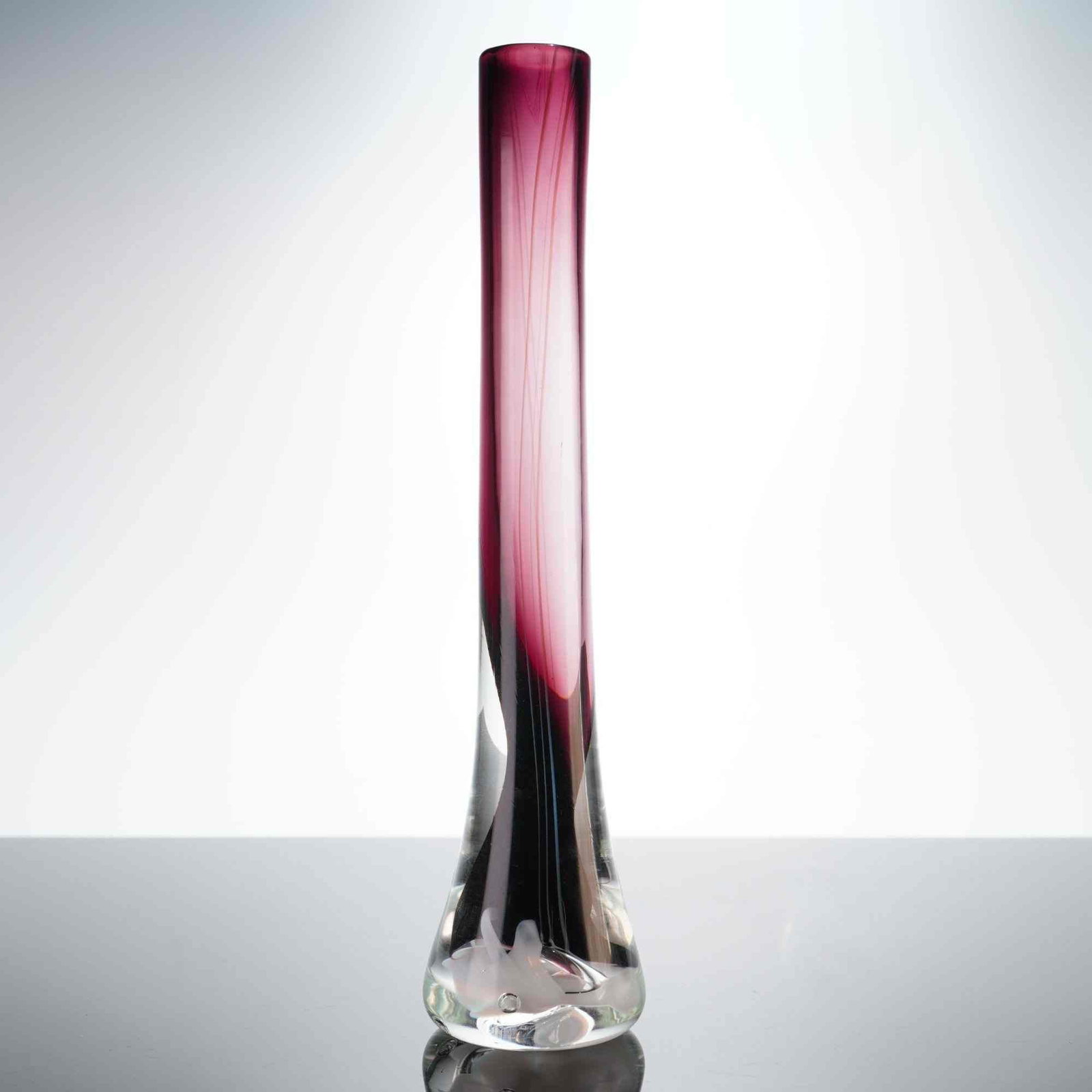 Richard Jolley Studio Art Glass Bud Vase: The piece is designed in a tall, slender tapering cylindrical form that transitions into a flared base. It is constructed using a casing technique, featuring a rich plum-colored interior core encased 