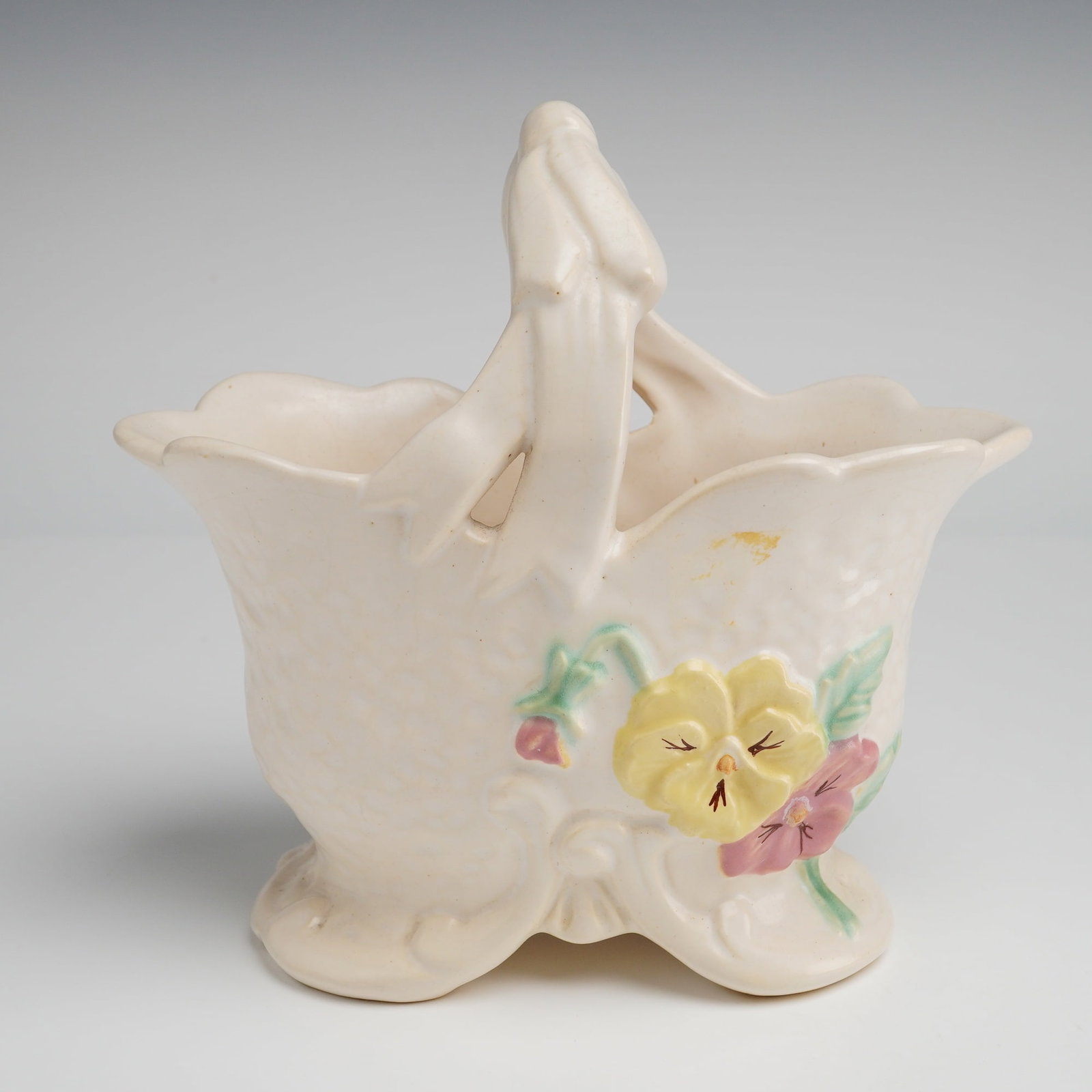 Weller Wild Rose Basket: This mid-century art pottery basket was produced by the Weller Pottery Company as part of the Wild Rose pattern. The glazed earthenware vessel features a high, central overhead handle topped with a mo