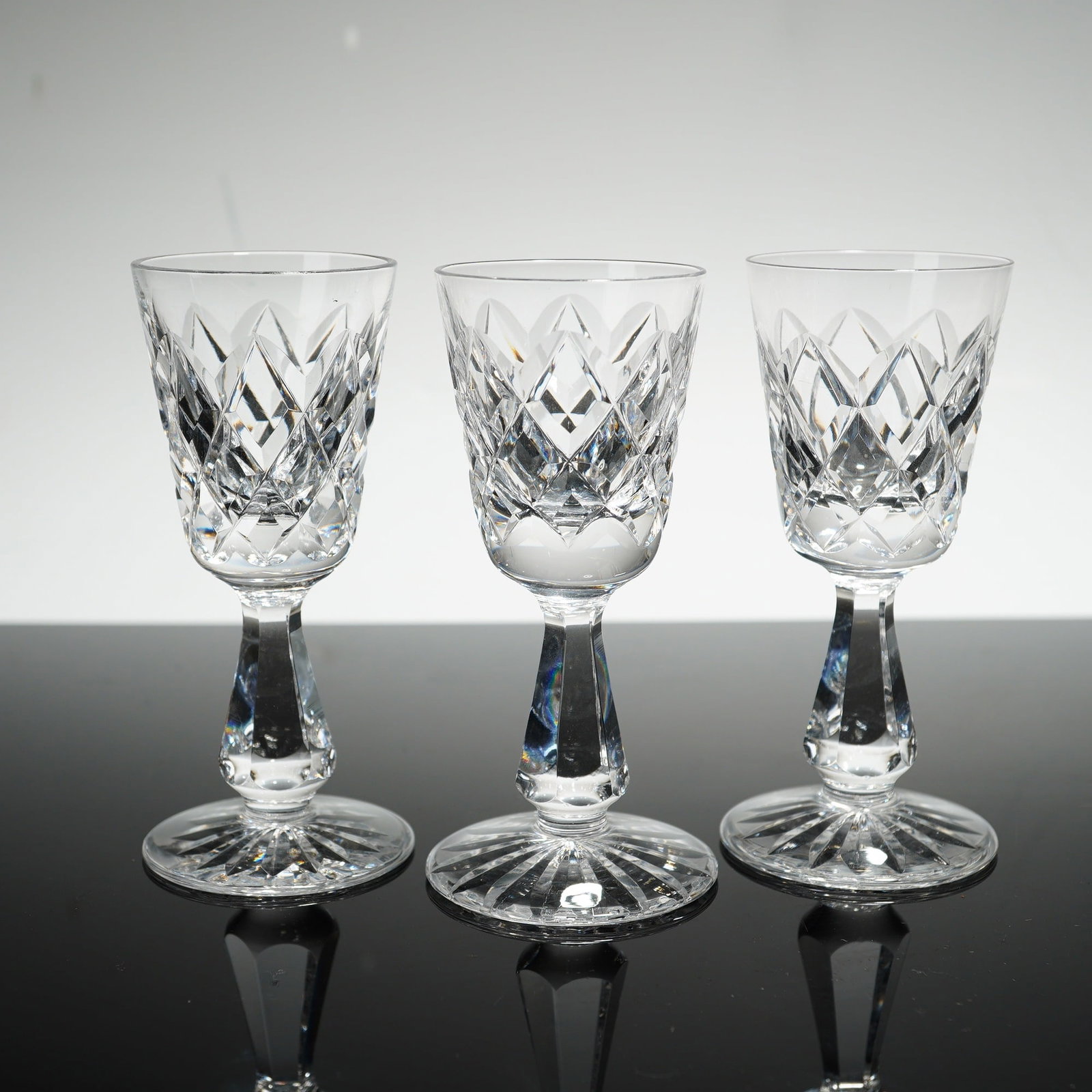 3pc Waterford Crystal Kinsale Cordial Glasses: The design showcases a sophisticated arrangement of deep-cut crisscross diamonds on the lower half of the bowl, positioned above a series of short vertical wedge cuts. The glasses feature a multi-face