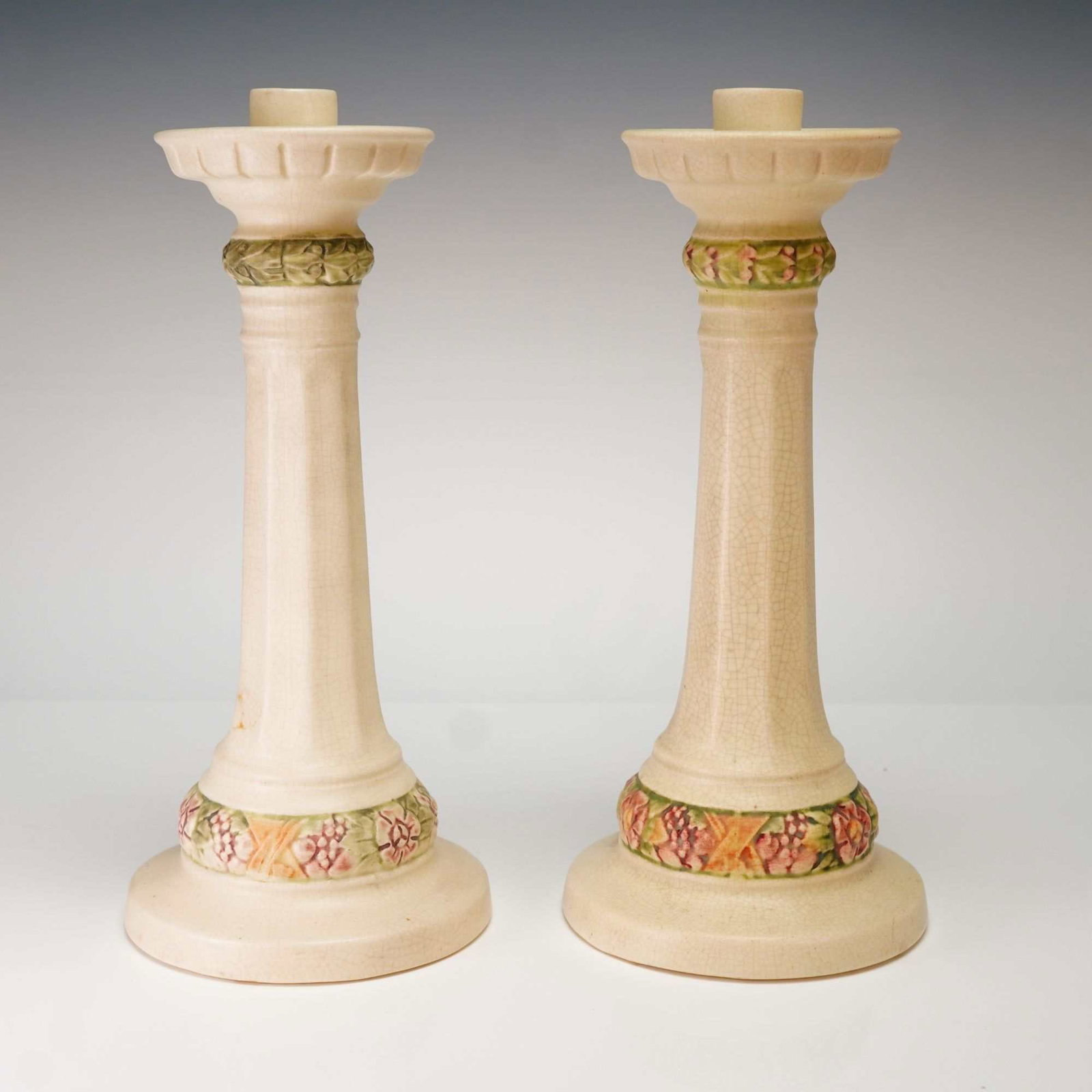 A Pair of Weller Pottery Roma Candlesticks: A pair of Weller Pottery ceramic candle holders from the Roma line. Each candlestick features a classical columnar form with a fluted stem rising from a flared base. The items are finished in a cream-