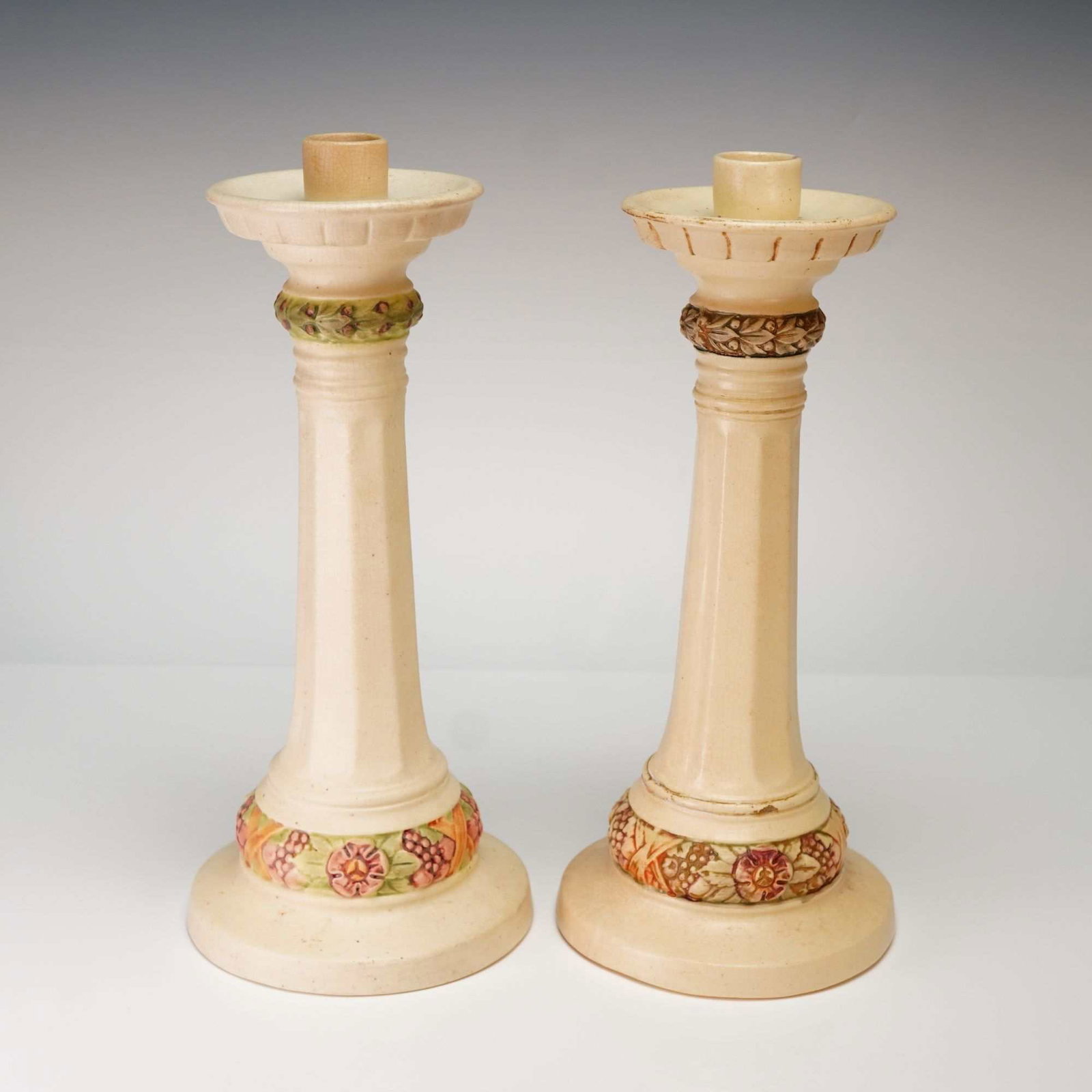 A Pair of Weller Pottery Roma Pattern Tall Candlesticks: A pair of Weller Pottery tall candle holders in the Roma pattern. These ceramic pieces feature a matte cream-colored glaze over a tall, tapered, and faceted shaft. Each candlestick rises from a broad,