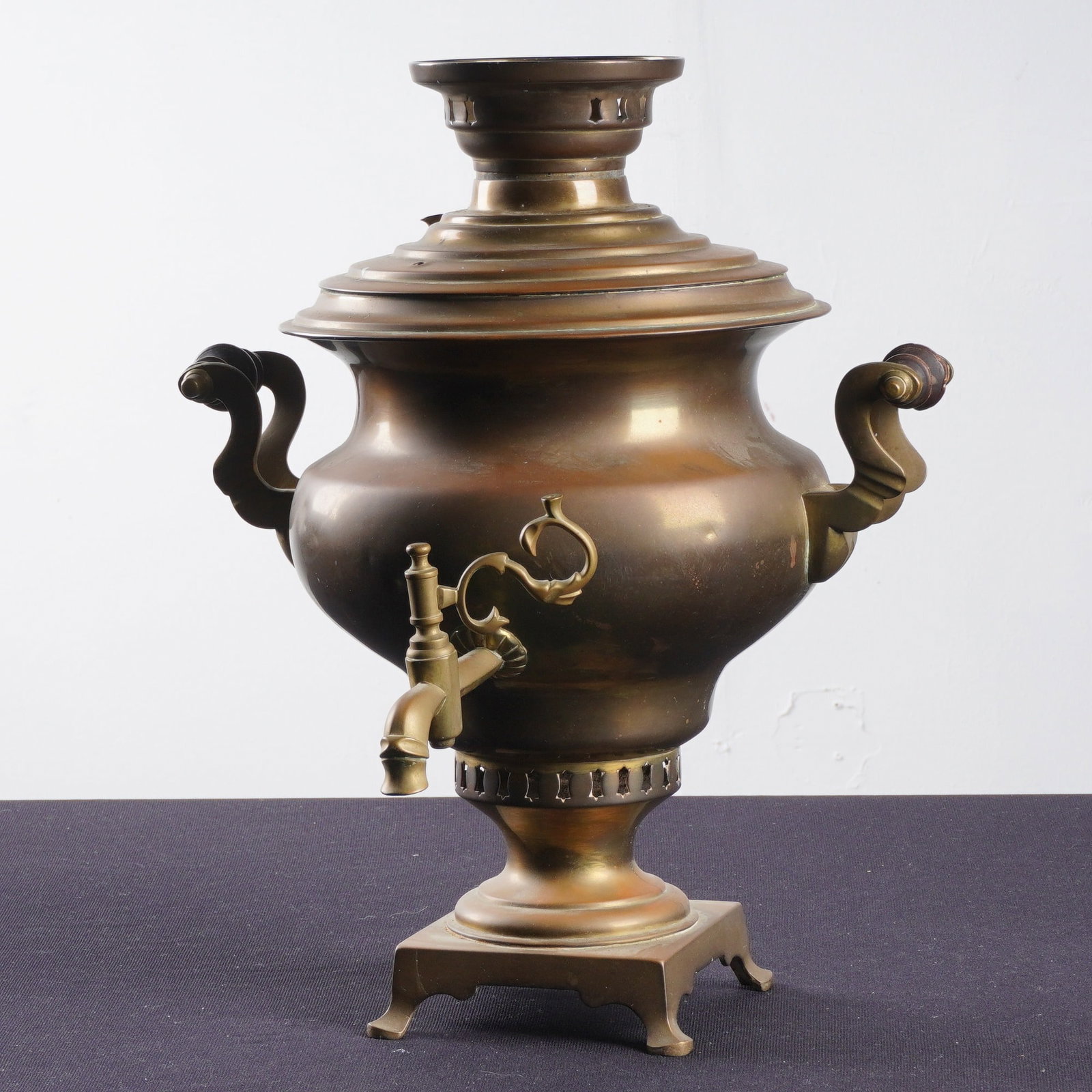 Antique N.A. Vorontsov Russian Brass Samovar: The construction features a spun brass body and lid with decorative cast brass elements including the scrollwork handles, spigot, and pedestal base. The piece reflects the traditional metalworking tec