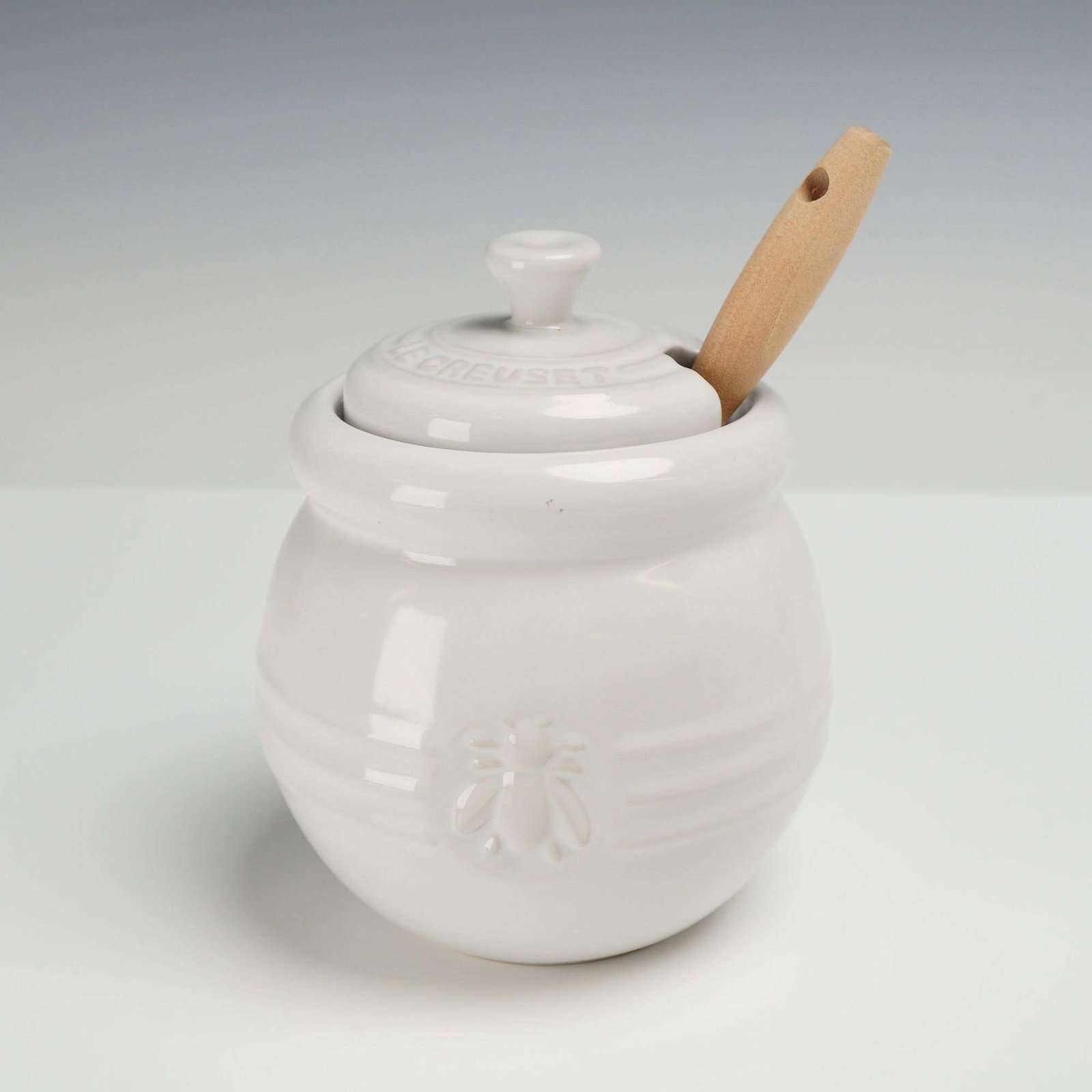 Le Creuset White Stoneware Honey Pot with Dipper: A 16 oz ceramic lidded honey pot with a wood and silicone dipper. The pot features an embossed honeybee on the front. The underside is marked Le Creuset and 16-24. Dimensions: 4"W x 5.75"H Weight: 13.