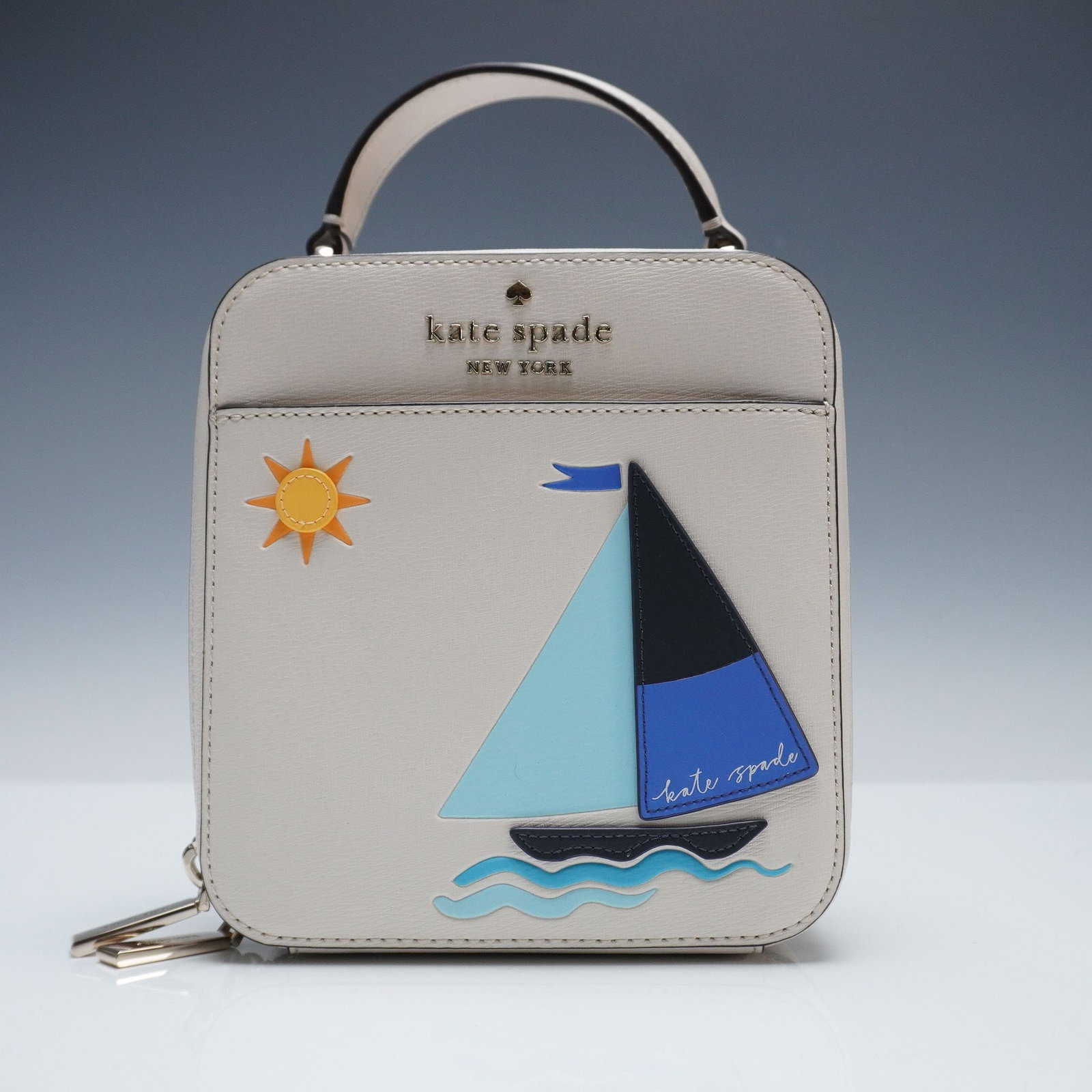 Kate Spade Sailboat Crossbody Vanity Bag: Charming and whimsical Kate Spade New York crossbody bag featuring a sailboat applique with sun and wave motifs on a structured cream-colored leather body. This compact handbag offers a top handle, du