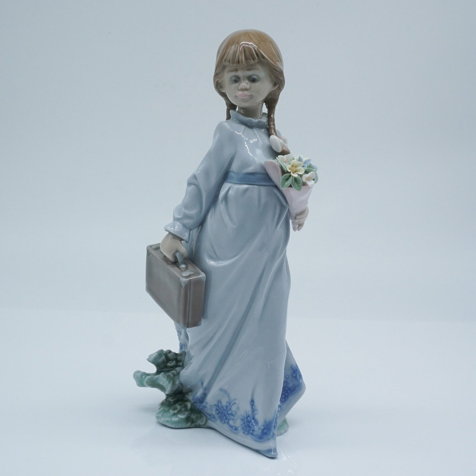 Lladro Porcelain Figurine, School Days 1007604: Charming Lladro porcelain figurine titled School Days (1007604), depicting a young girl with braided hair holding a bouquet and a suitcase, rendered in soft pastel tones with delicate floral detail, t