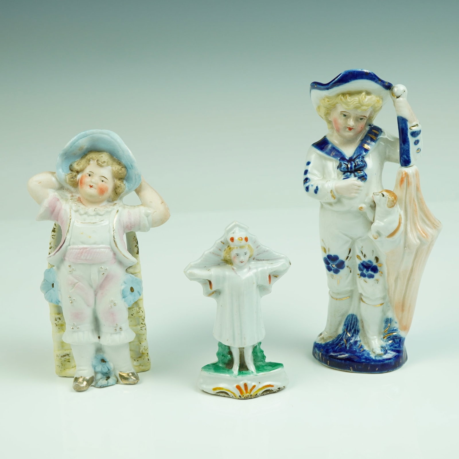 Porcelain Children Figurines: An utterly charming trio of figural fairings depicting children in whimsical poses, each crafted with delightful detail and hand-painted charm. The group includes a young boy in a pastel pink suit wit