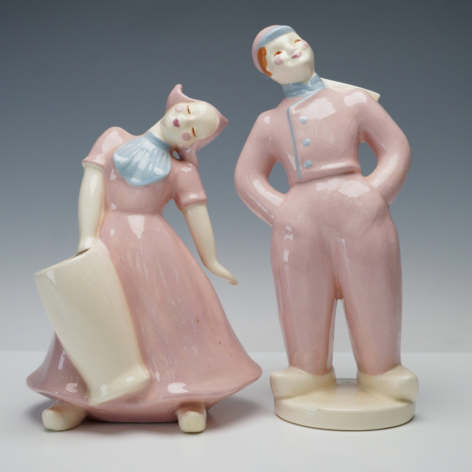 Pair of Heidi Schoop Art Creations Ceramic Figurines, Dutch Boy and Girl: Each figure is hand-painted in soft pastel pink with light blue accents. The boy is shown standing with hands on hips, wearing a cap, scarf, and trousers, while the girl wears a traditional dress and 