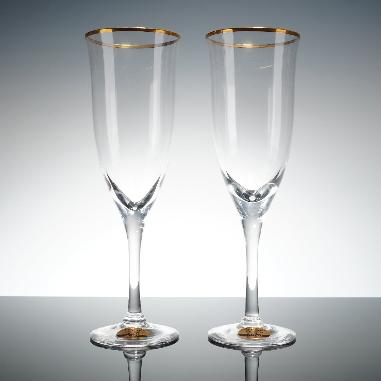 Pair of Noritake Crystal Champagne Flutes, Troy: The flutes are constructed from full lead crystal and feature elongated clear bowls accented by a gilded gold rim. Each glass is characterized by a tapered silhouette set upon a slender stem and a fla