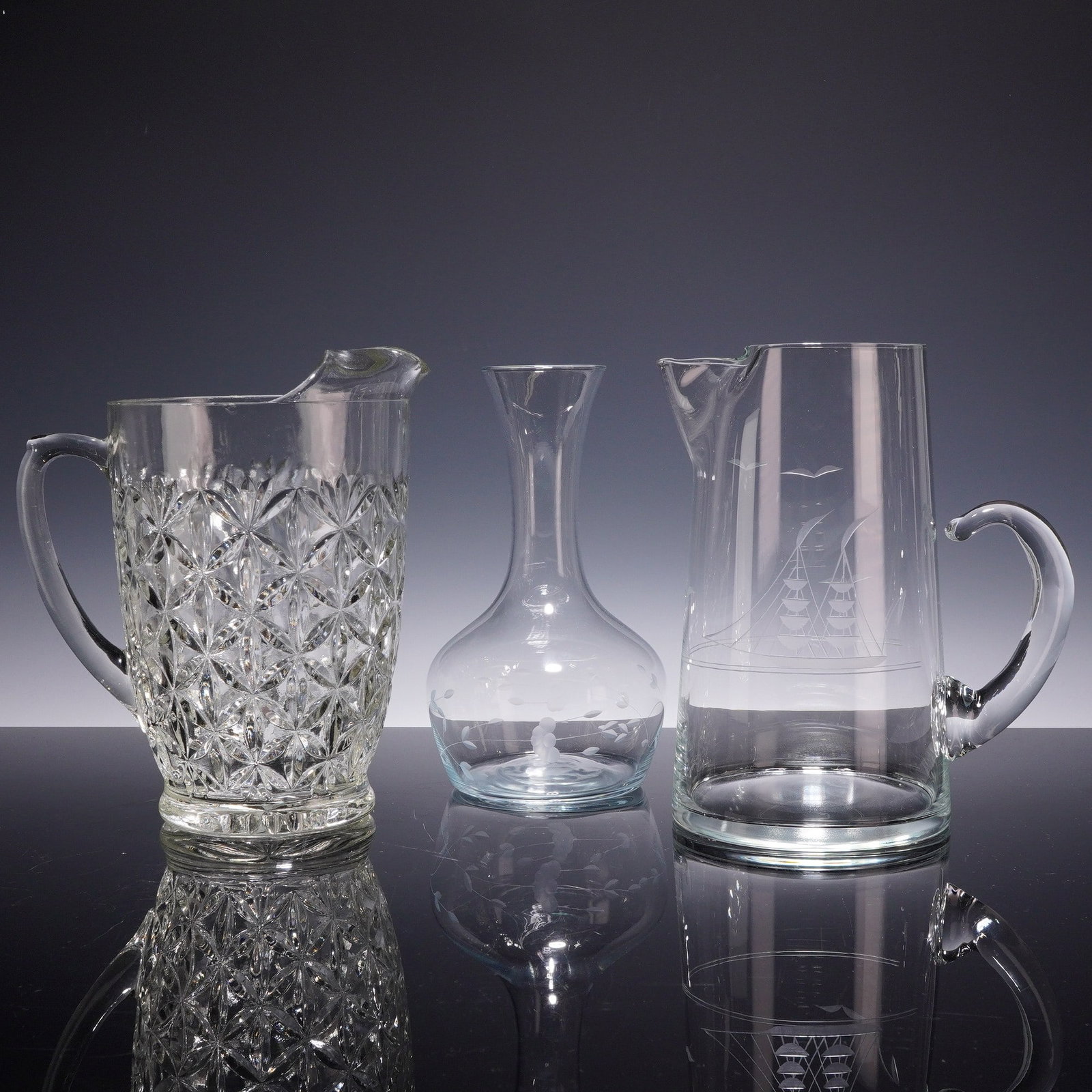 Three Clear Glass Items Including a Nautical Theme Pitcher, a Cut Pattern Pitcher, and a Carafe: This lot features three pieces of clear glassware. The first is a large handled pitcher decorated with a geometric cut pattern of repeating faceted diamonds and rosettes. It features a prominent pouri