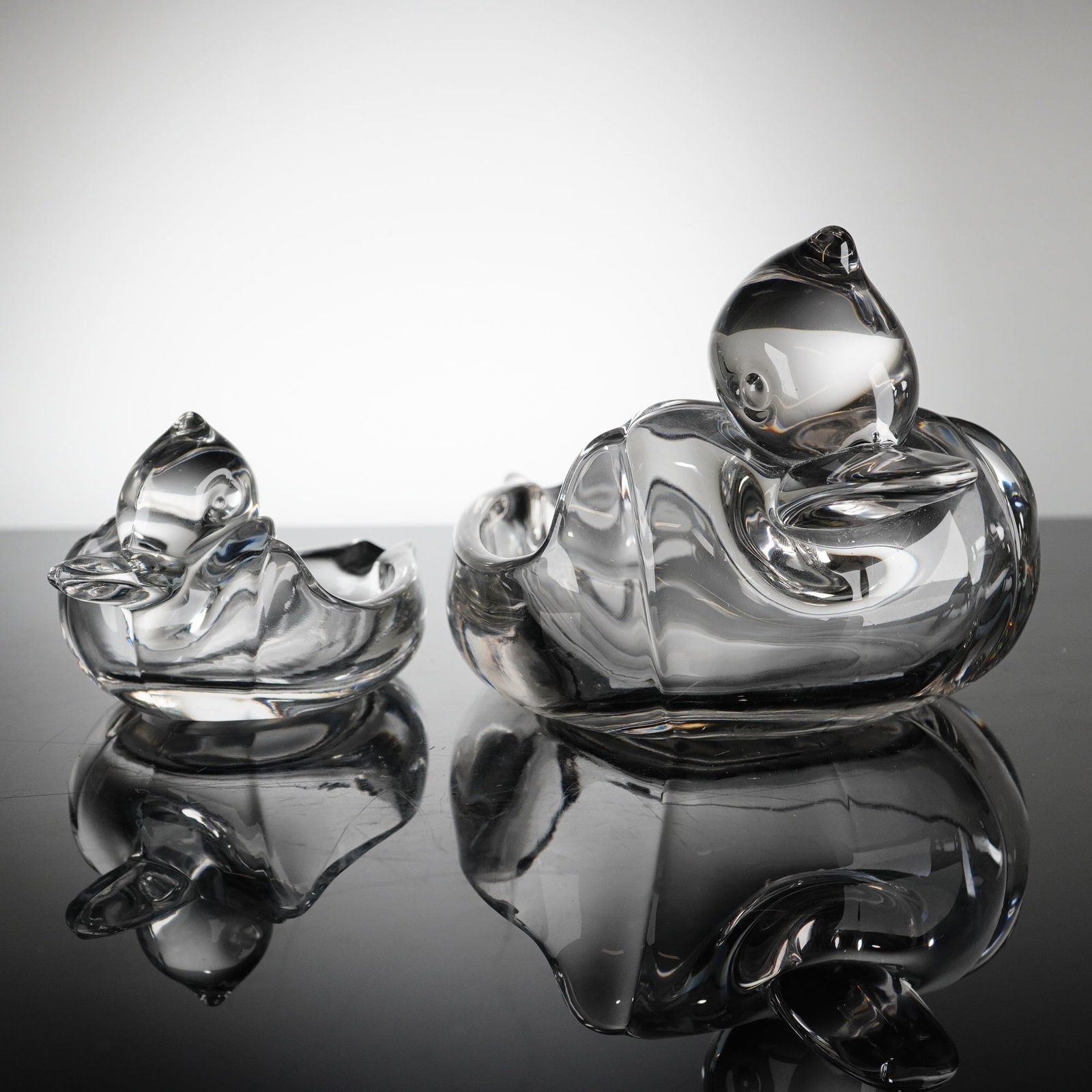 A Pair of Duncan & Miller Clear Glass Duck Ashtrays: A pair of heavy clear glass vessels attributed to Duncan & Miller, commonly known as the Pall Mall Glass Duck Ashtray. The items are molded and stylized in the form of a swimming aquatic bird, featuri