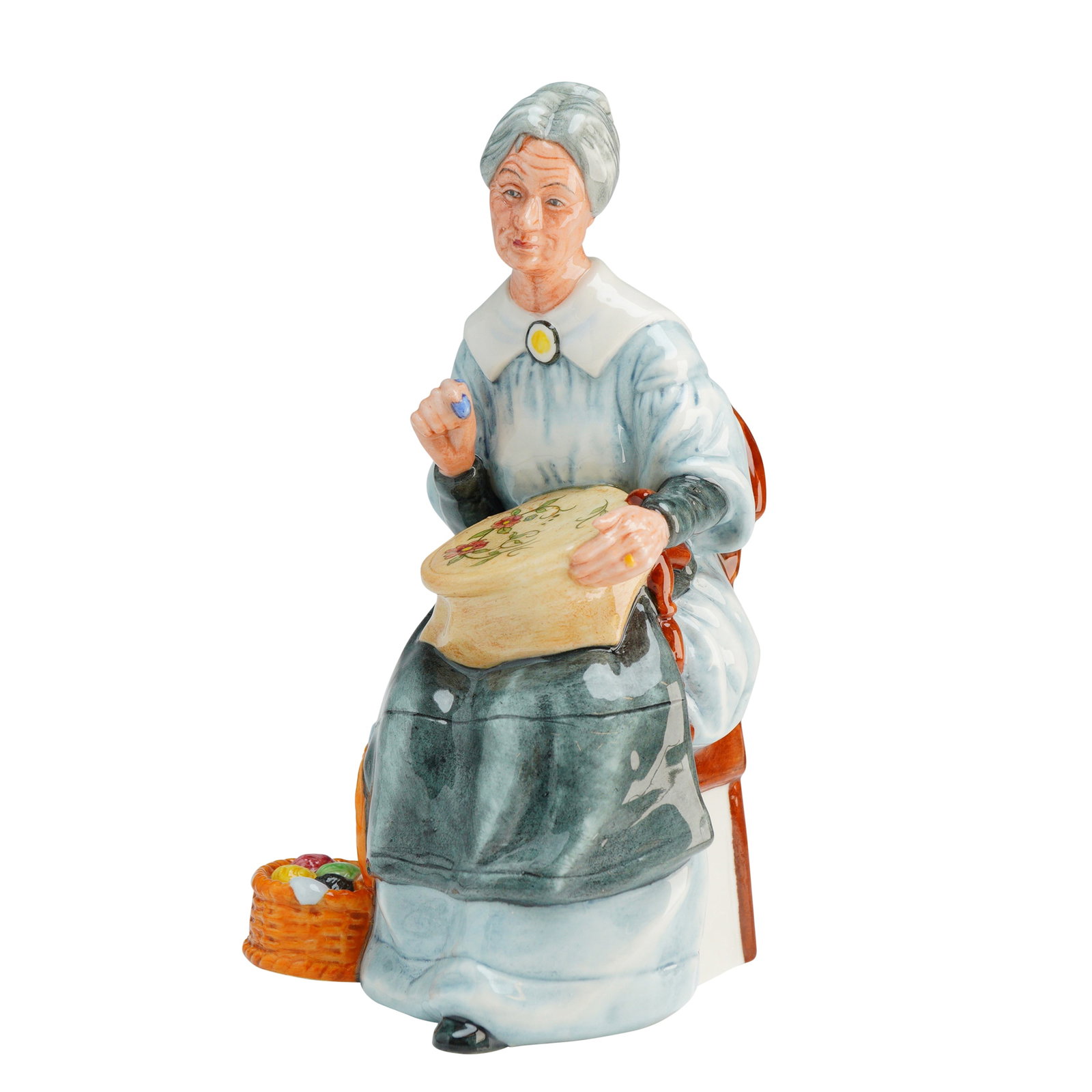 Royal Doulton Figurine, Embroidering HN2855: The figure depicts an elderly woman with grey hair styled in a bun, seated on a brown high-backed chair featuring green upholstery. She is dressed in a pale blue gown with a white collar and a dark gr