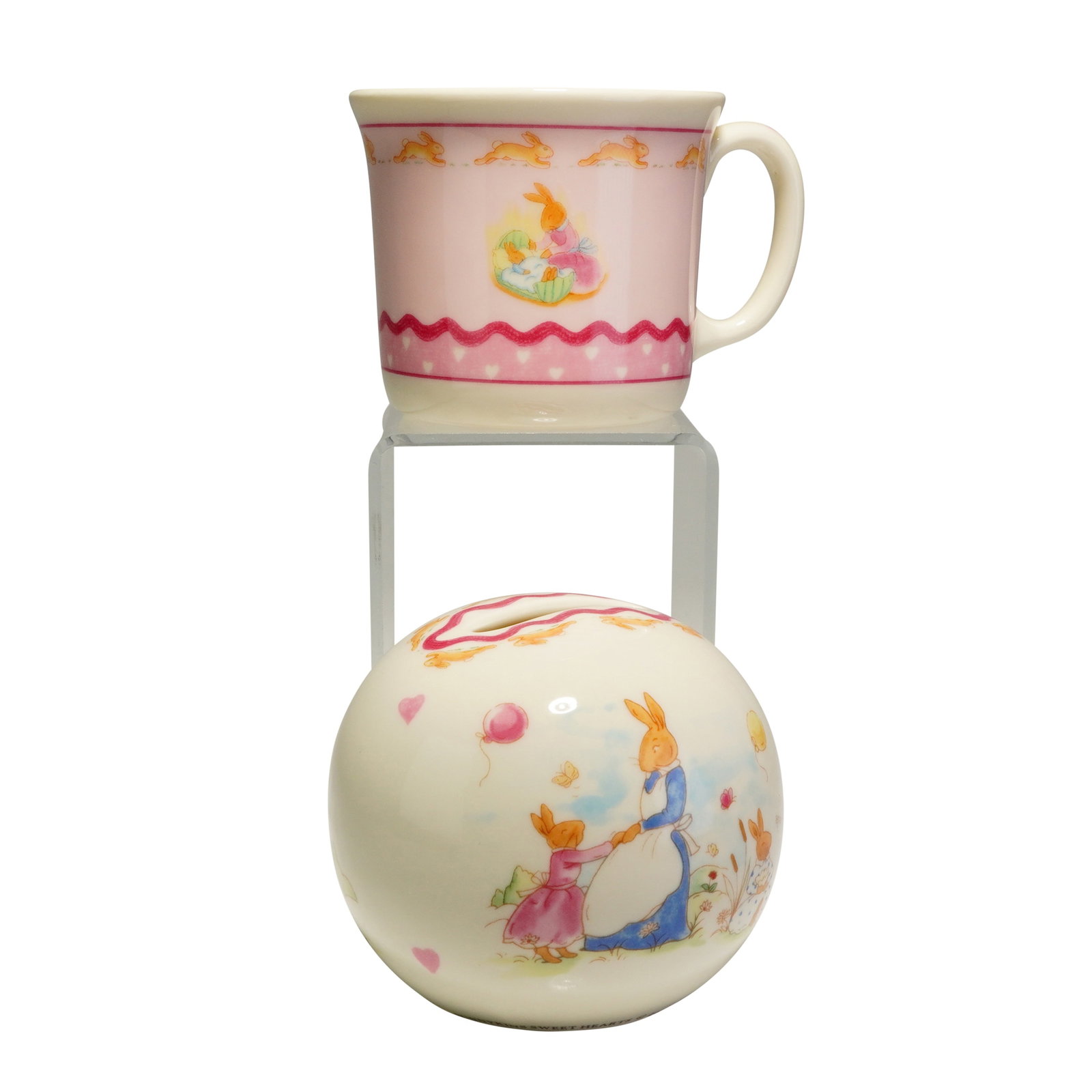 2pc Royal Doulton Bunnykins Sweet Hearts Mug and Money Bank: The handled mug and spherical money box are crafted from bone china with a cream-colored ground, while the mug features a prominent pink area as its main background. Both pieces showcase colorful illu