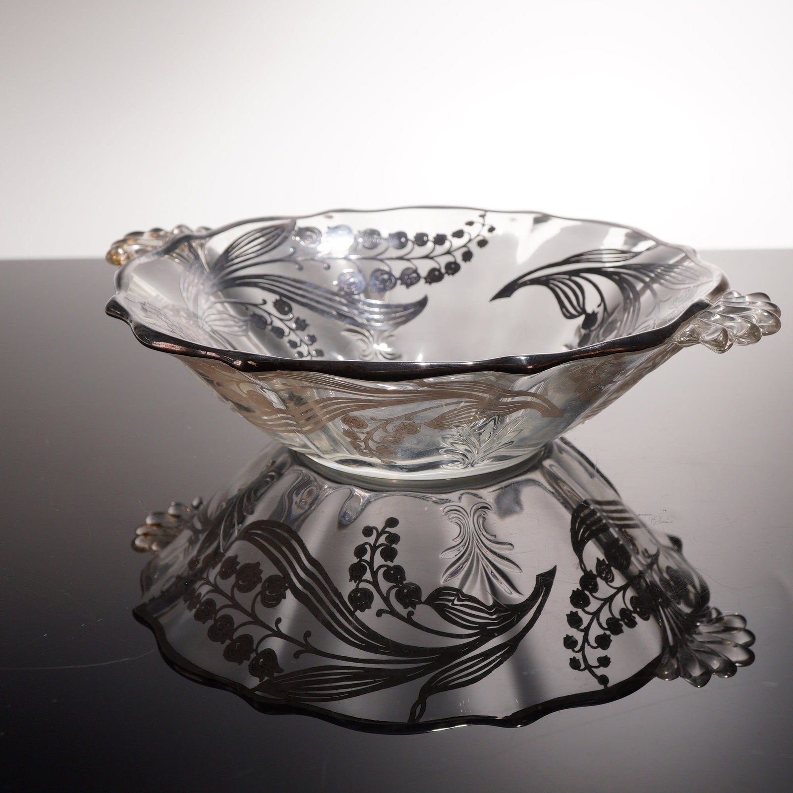 Sterling Silver Overlay Lily of the Valley Motif Handled Bowl: A handled bowl crafted from clear molded glass featuring intricate silver overlay in a Lily of the Valley motif. The circular form is characterized by a scalloped rim and two integral fan-shaped handl