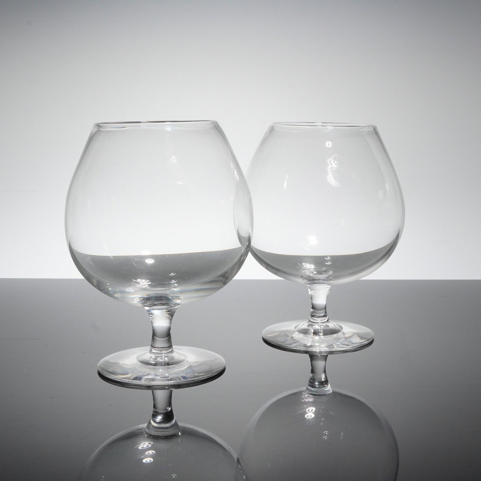 A Pair of Steuben Contemporary Clear Glass Brandy Snifter Glasses, Signed: A pair of brandy glasses, styled as contemporary snifters or balloons. Each glass features a large, globular bowl crafted from clear, highly reflective glass, supported by a short, thick baluster stem