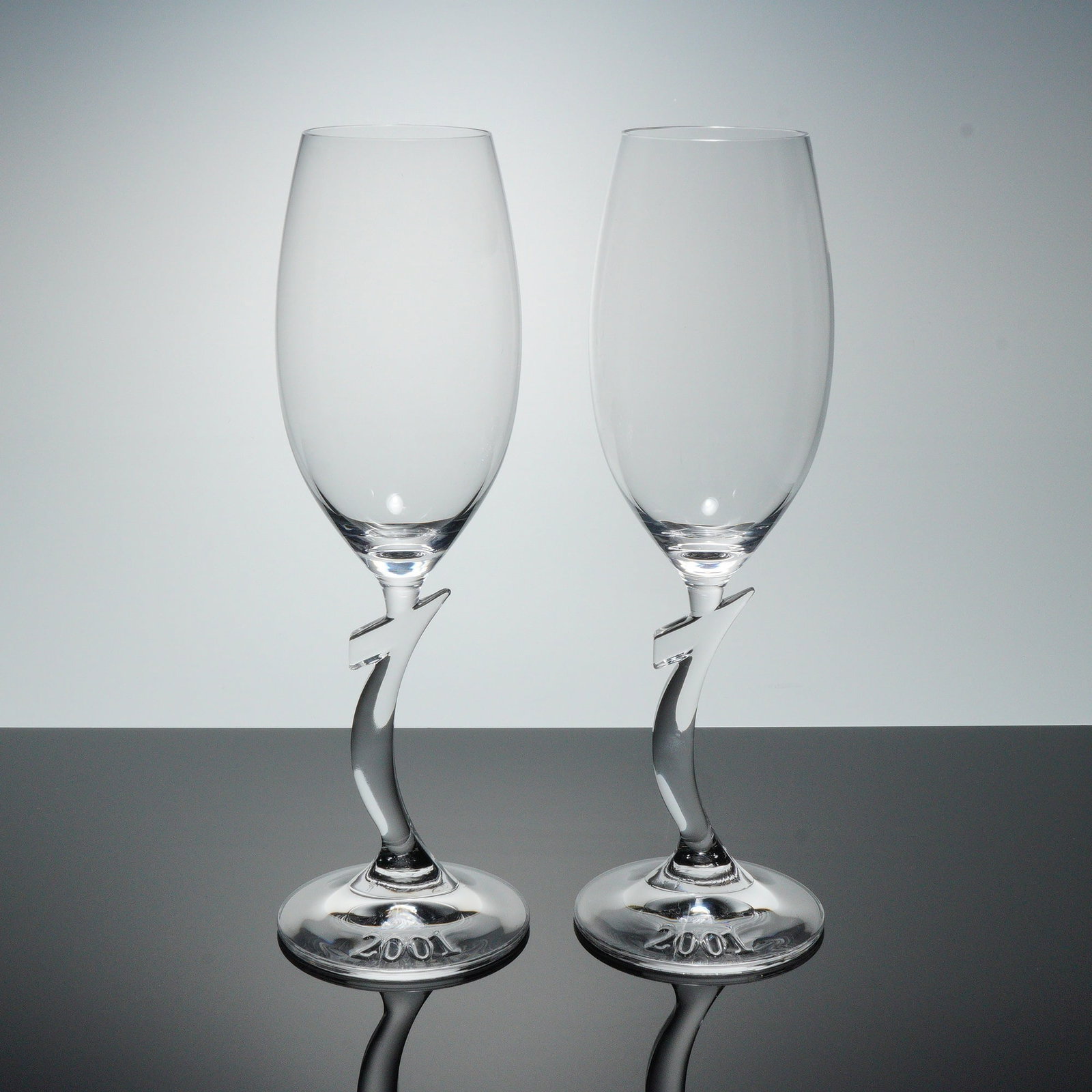 2pc Riedel Crystal Champagne Glasses, Millenium: A pair of Riedel crystal champagne glasses featuring a distinctive sculpted stem with a 1 form and the year 2001 etched to the foot, each signed Riedel, crafted in clear crystal Dimensions: Each glass