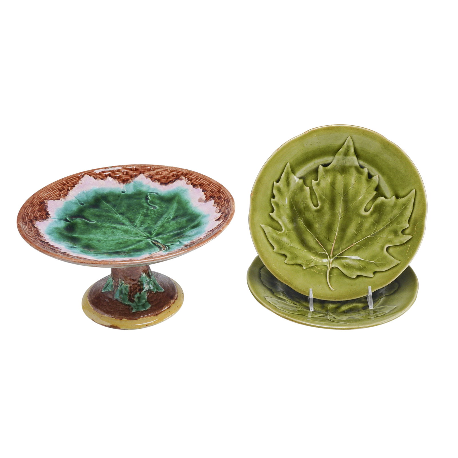 3pc H.B. & Cie and Griffen, Smith & Hill Majolica Compote and Plates: The grouping includes a compote featuring a vibrant green molded leaf center, a pink transition border, and a brown basketweave textured rim. The compote is supported by a flared pedestal base decorat