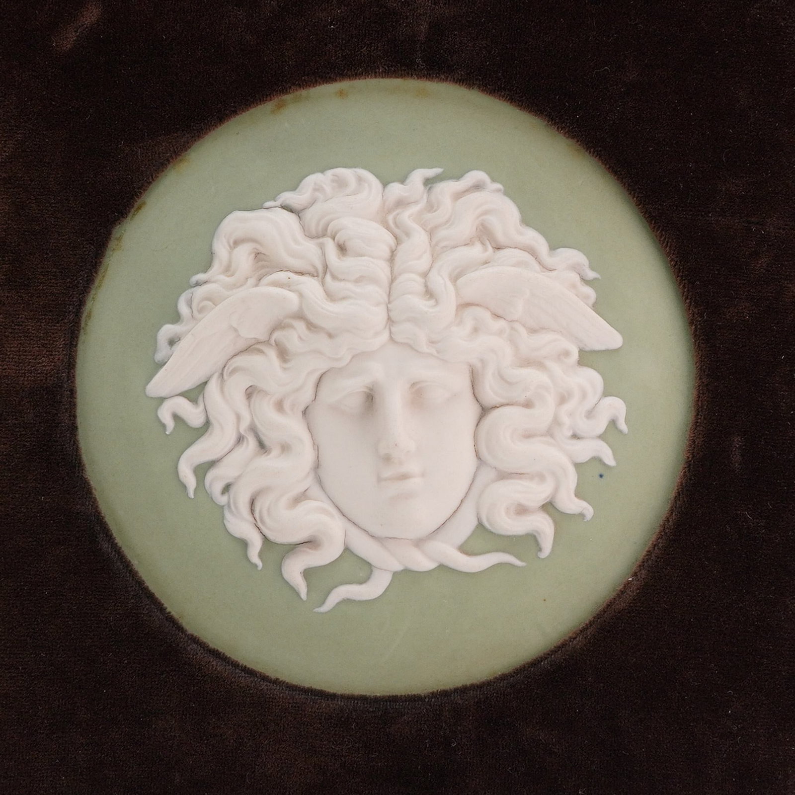 Wedgwood Sage Green Jasperware Medusa Plaque: A circular Wedgwood jasperware plaque featuring a high-relief white sprigged decoration of the head of Medusa, modeled after the Medusa Rondanini. The goddess is depicted in a frontal view with wings 