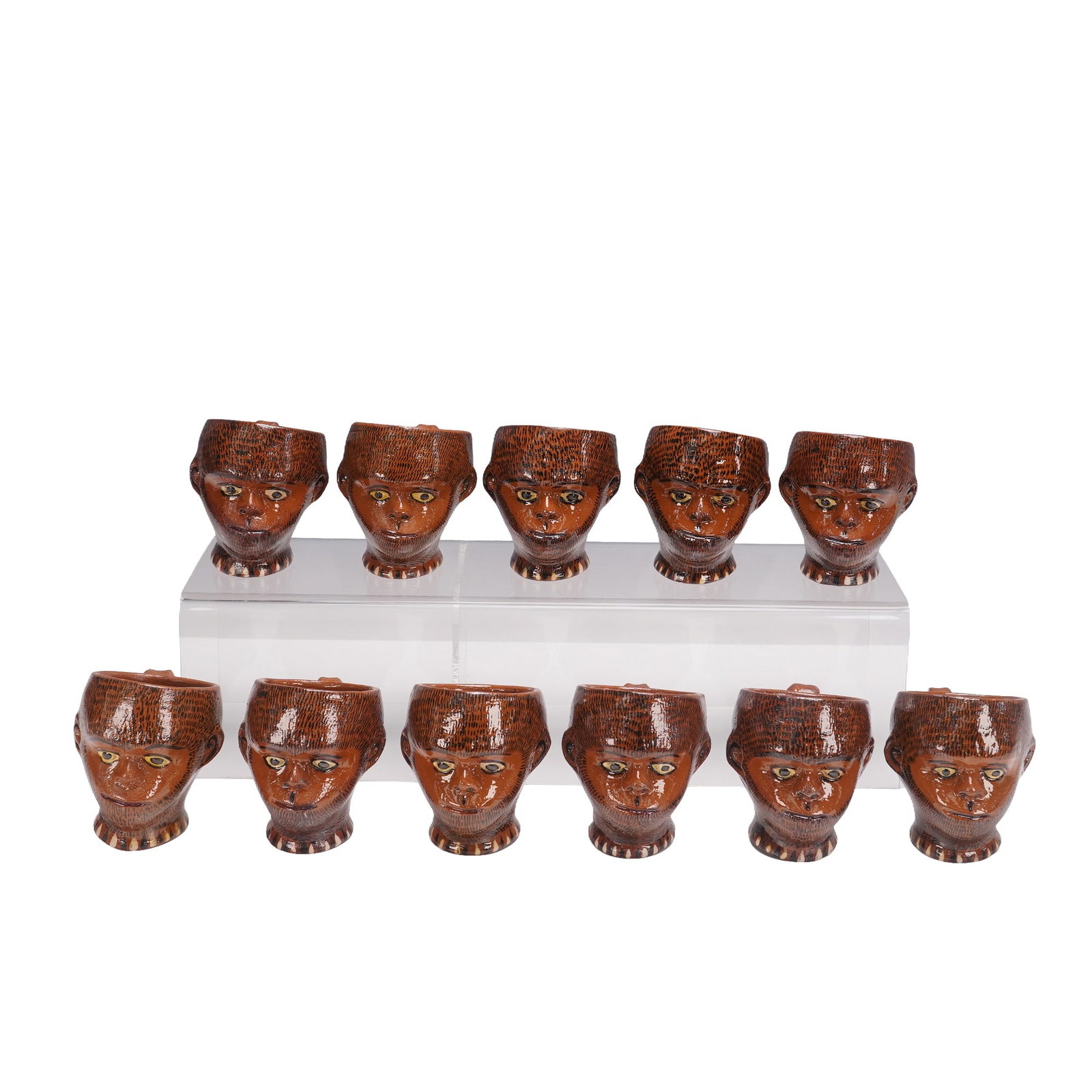 11pc Vintage Hand-Painted Character Jugs: A set of eleven small ceramic jugs, each modeled in the form of a stylized head with detailed facial features, including prominent eyes, brows, and textured hair. The jugs are hand-painted in warm bro
