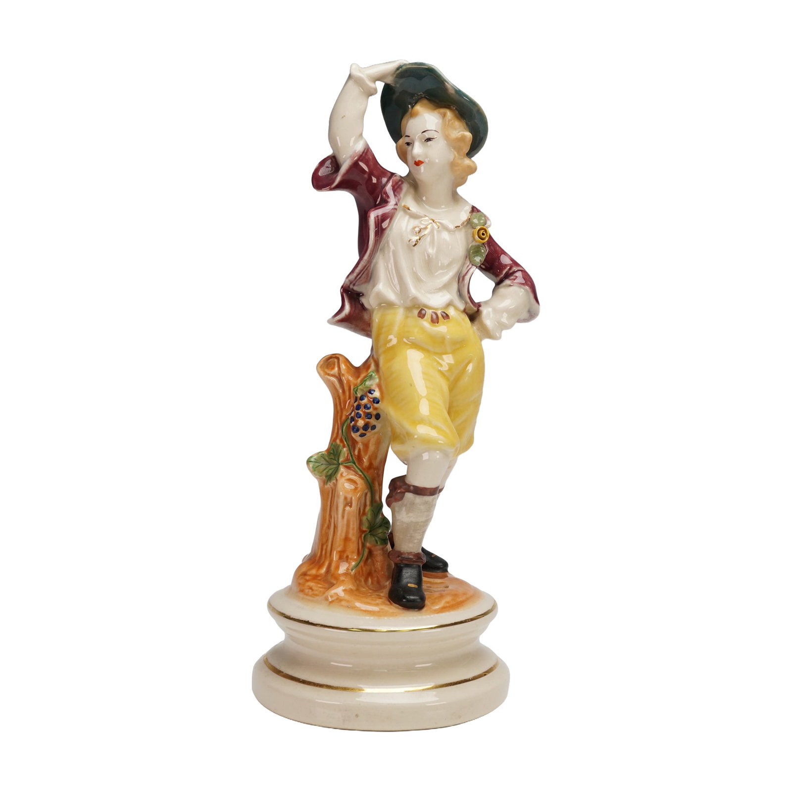 Hand-Painted Porcelain Country Boy Figurine: Hand painted porcelain figure of a carefree young man in yellow breeches and green hat, posed beside a tree stump with applied grape and leaf detail, raised on a circular stepped base with gilt line. 