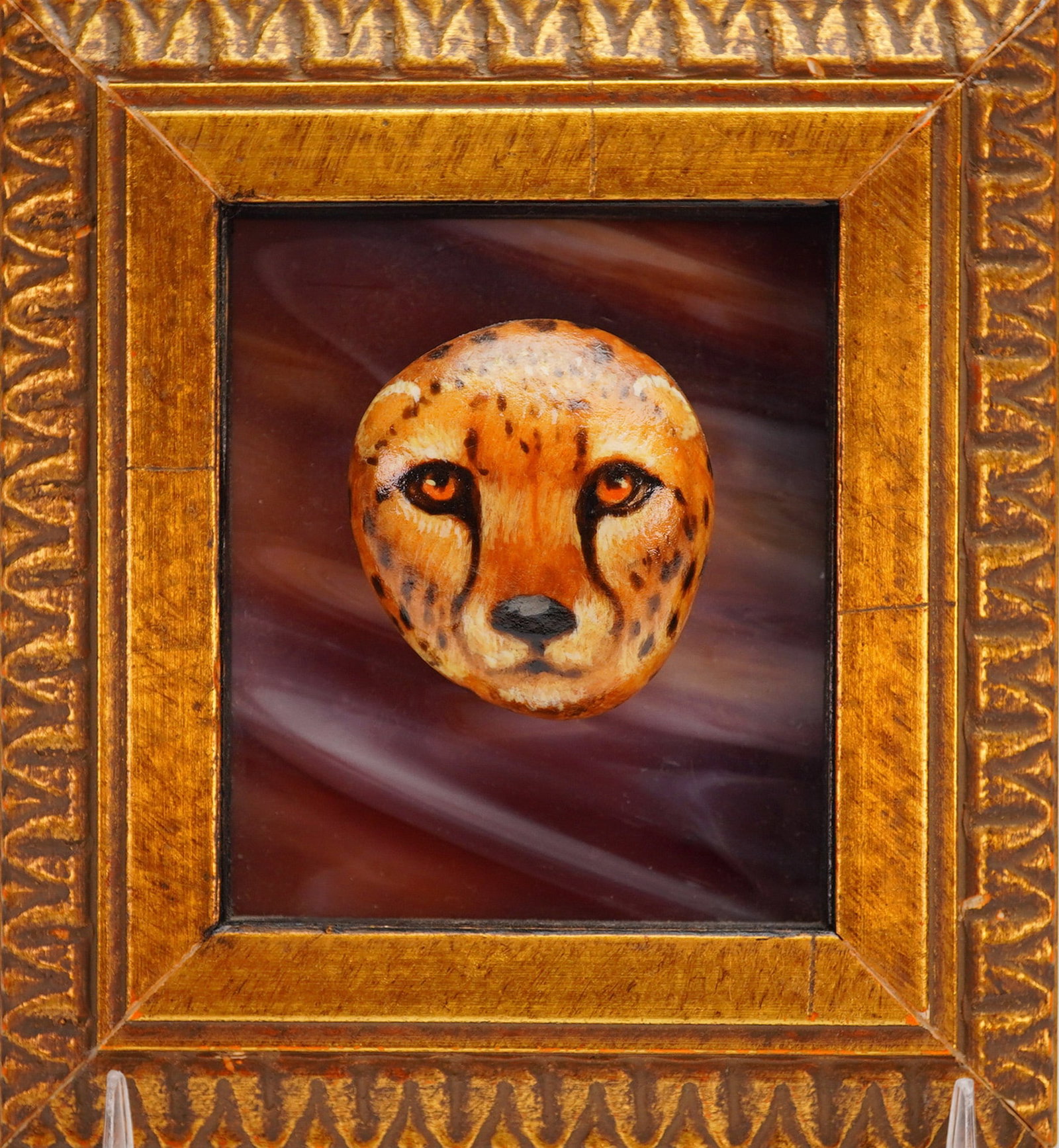 Framed David Homan Painted Plaque, Rock Cheetah: David Homan. A miniature sculptural relief depicting the head of a cheetah, featuring painted details, bright orange eyes, and spotted fur. The relief is mounted against a swirled, reddish-purple back