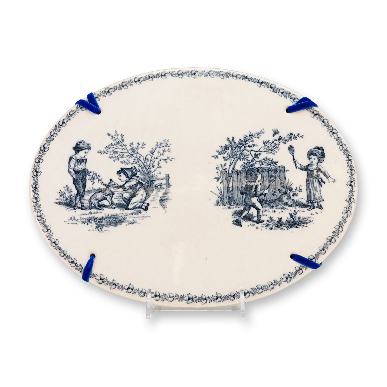 French Transfer-Printed Ceramic Decorative Hanging Wall Plate from the Rubin Collection: A French transfer-printed earthenware plaque of oval form, dating to approximately 1880. The cream-toned ground features two distinct blue vignettes depicting children in period attire engaged in outd