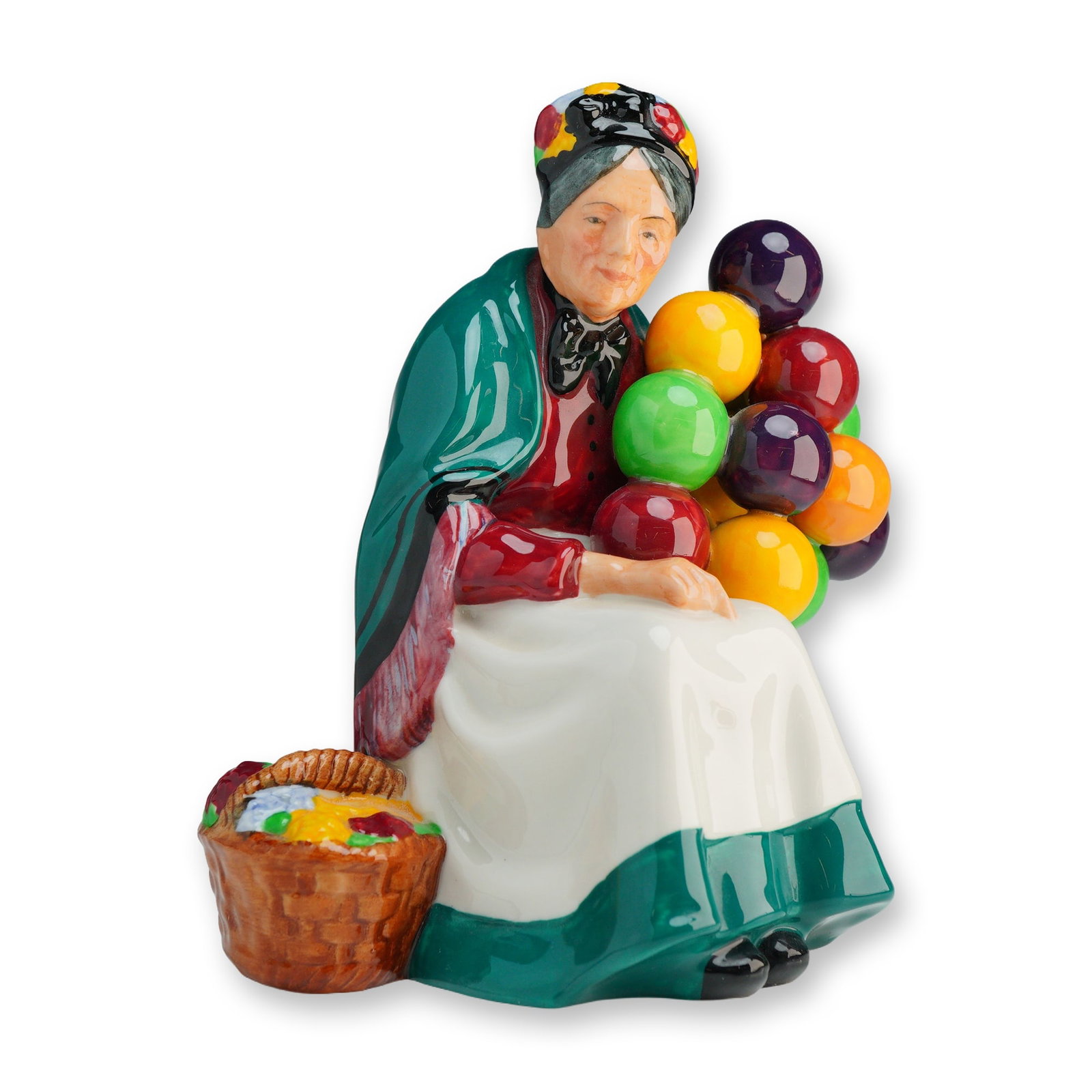 Royal Doulton Figurine, Old Balloon Seller HN1315: The figure depicts an elderly woman seated on a brown crate. She is dressed in a green shawl over a red bodice and a white apron with a dark green border, with a patterned headscarf tied over her grey