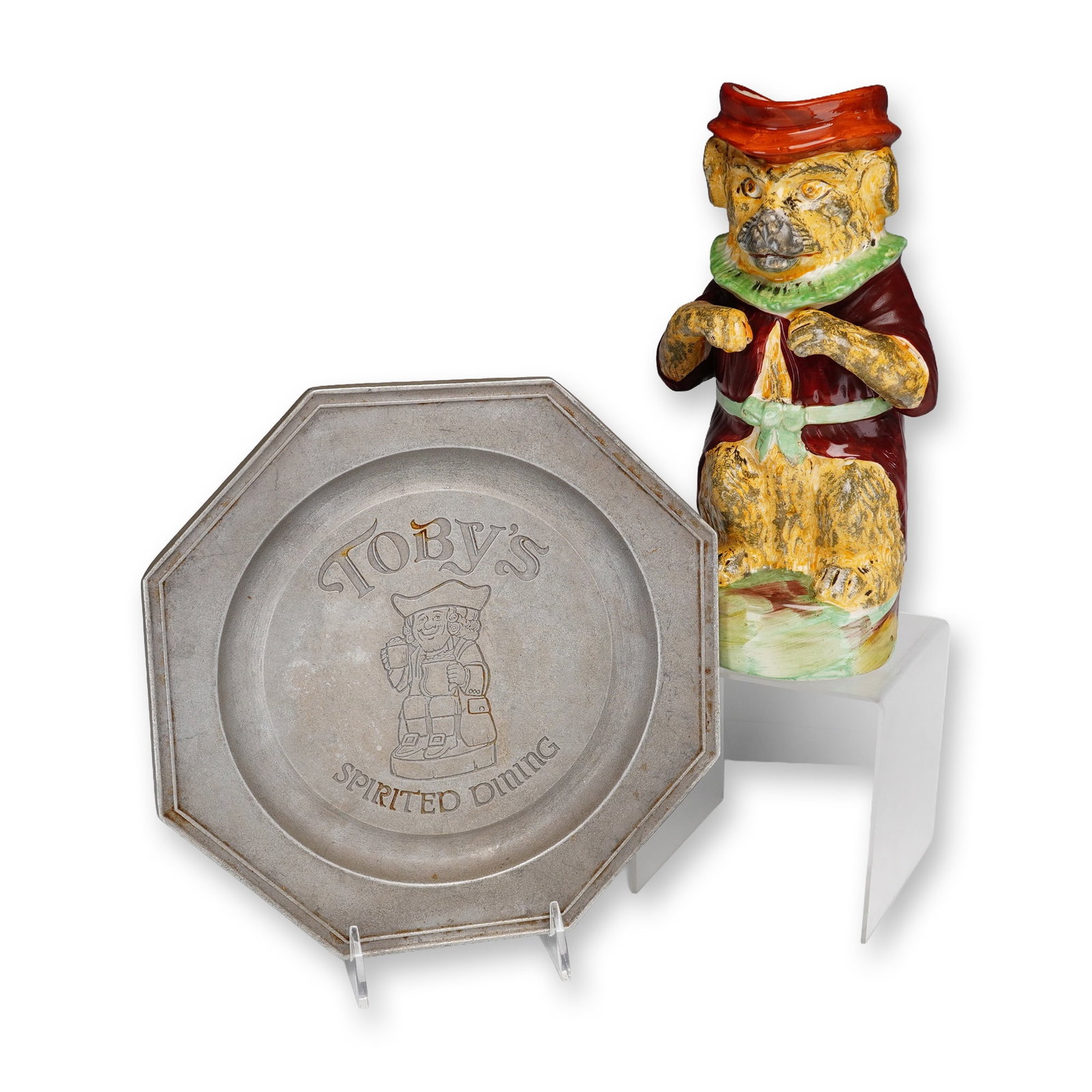 Two-Piece Lot Featuring a Dog Figural Toby Jug and a Wilton Armetale Plate: This lot consists of a figural ceramic Toby jug and a commemorative metal plate. The pitcher is modeled as a seated dog wearing a red tricorn hat, a burgundy coat, and a green ruff collar. The handle 