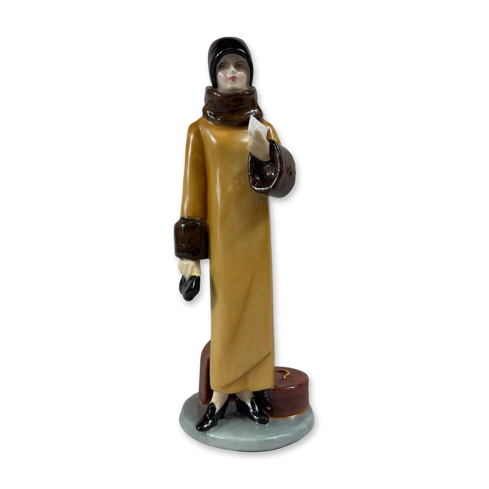 Royal Doulton Bone China Figurine, Waiting for a Train HN3315: The piece depicts a woman in stylized early 20th-century attire, wearing a mustard-yellow coat with dark brown fur-effect trim at the collar and cuffs, a black hat, and black heeled shoes. The figure 