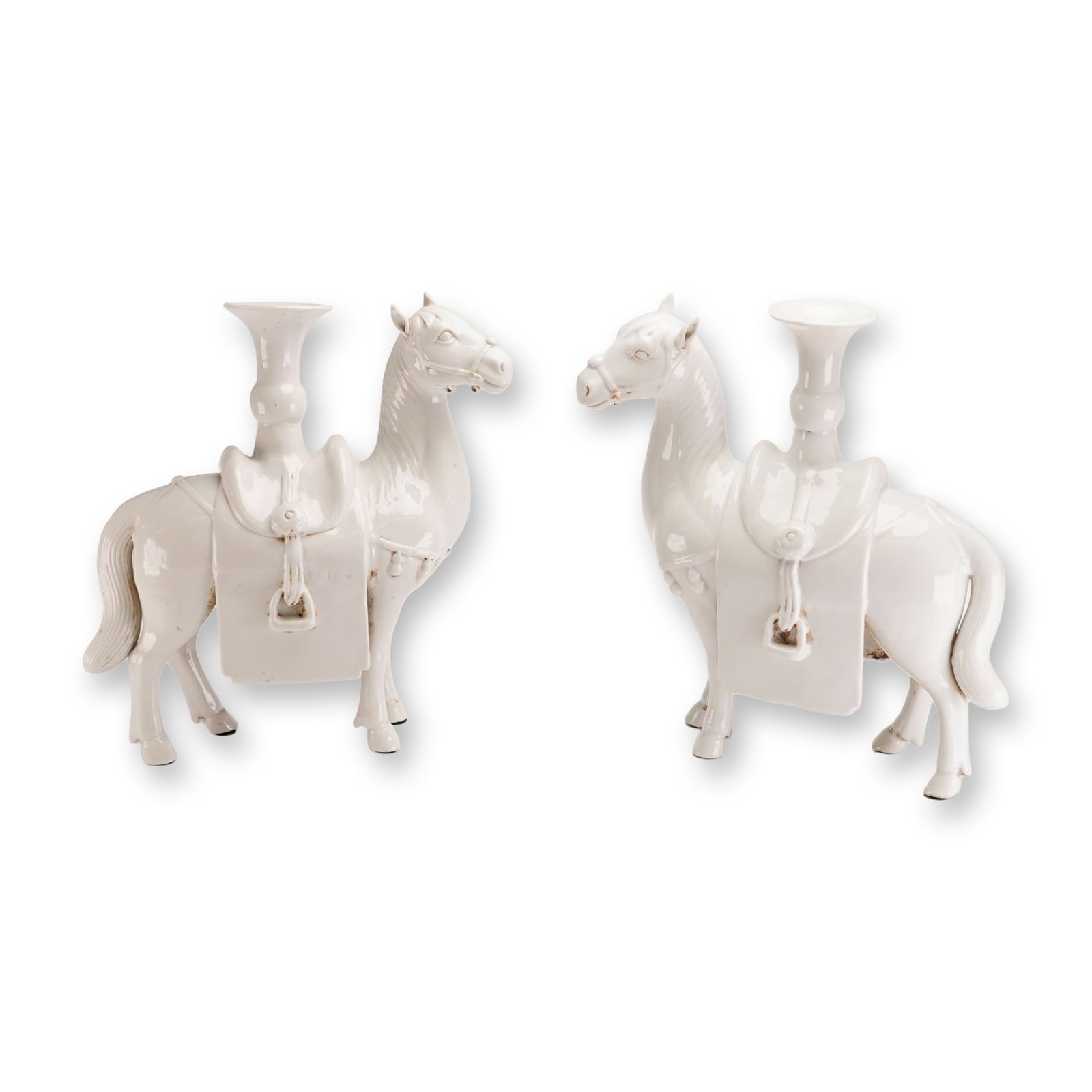 Pair of White Glazed Ceramic Figural Horse Candle Holders: Each horse is modeled in a standing position with the head turned to the side, featuring detailed molded trappings including bridles, tasseled breastplates, and saddles. A trumpet-shaped vessel with a
