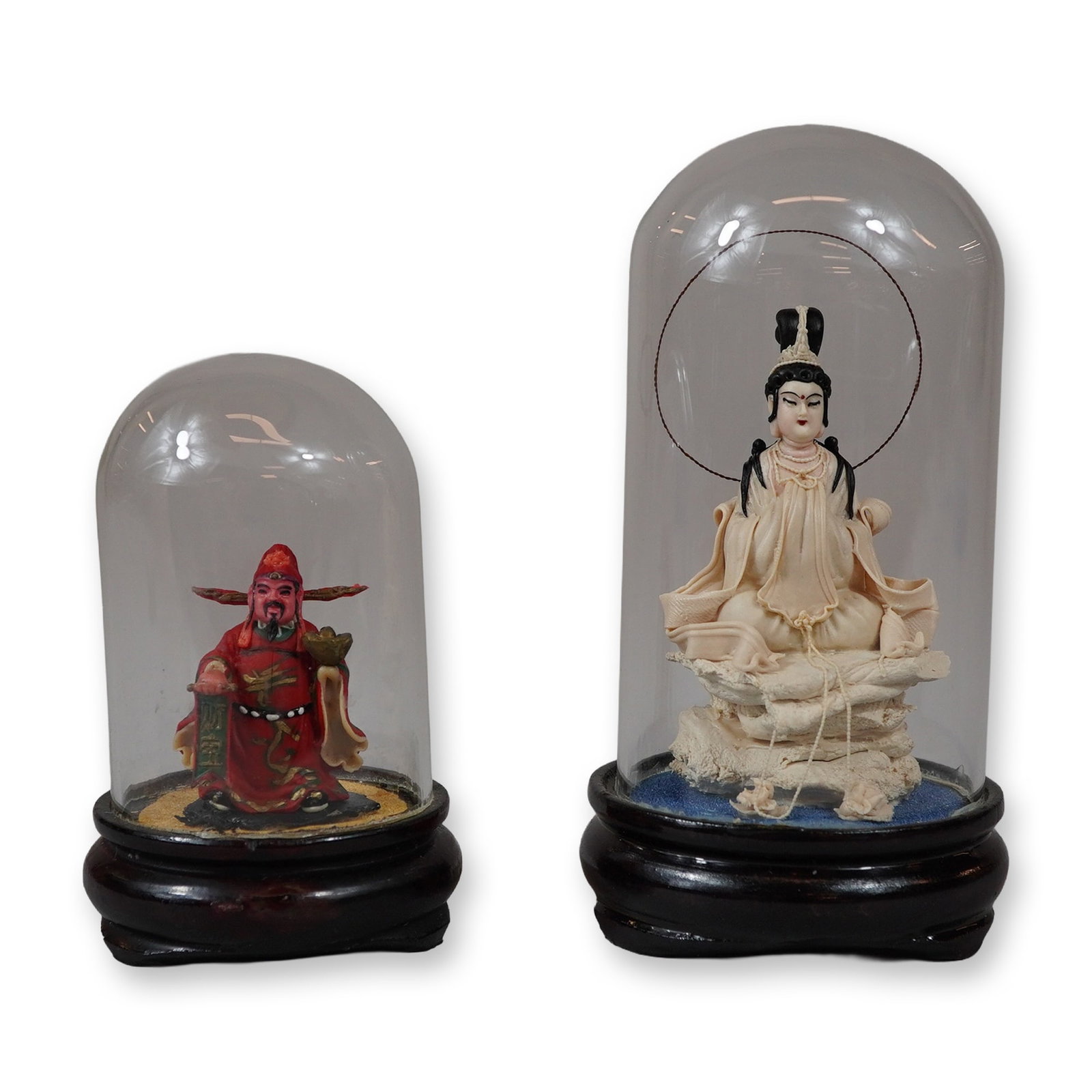 Two Chinese-Style Decorative Figurines Under Glass Cloches: This lot features two miniature Chinese-style decorative figurines, each housed under a glass cloche on a turned ebonized wood base. The smaller figure represents a male deity, likely Caishen, wearing