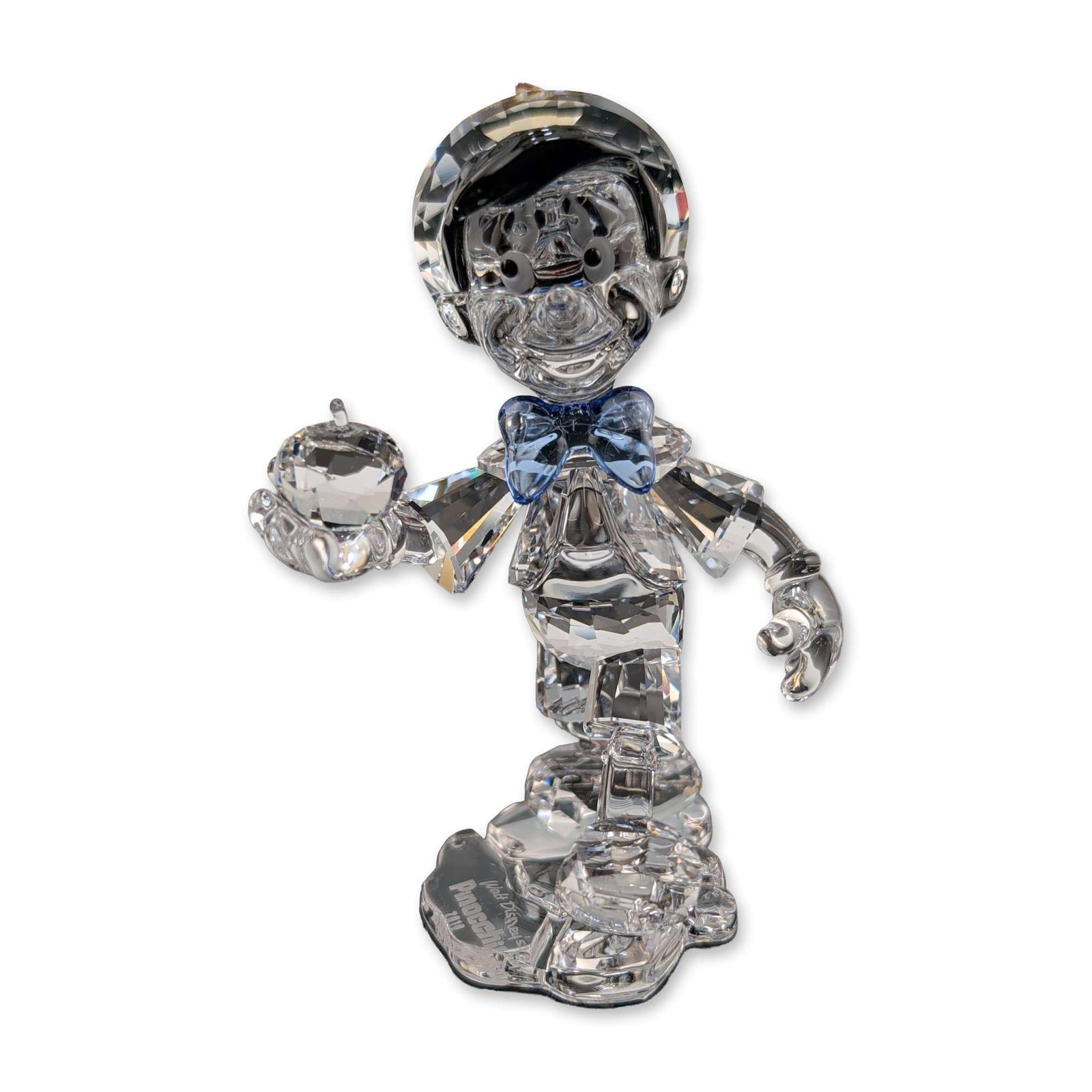 Swarovski Disney Crystal Figurine, Pinocchio: Swarovski Disney character figurine in clear and colored faceted crystal, depicting Pinocchio in a walking pose while holding an apple. The figure features a clear faceted body with a blue crystal bow