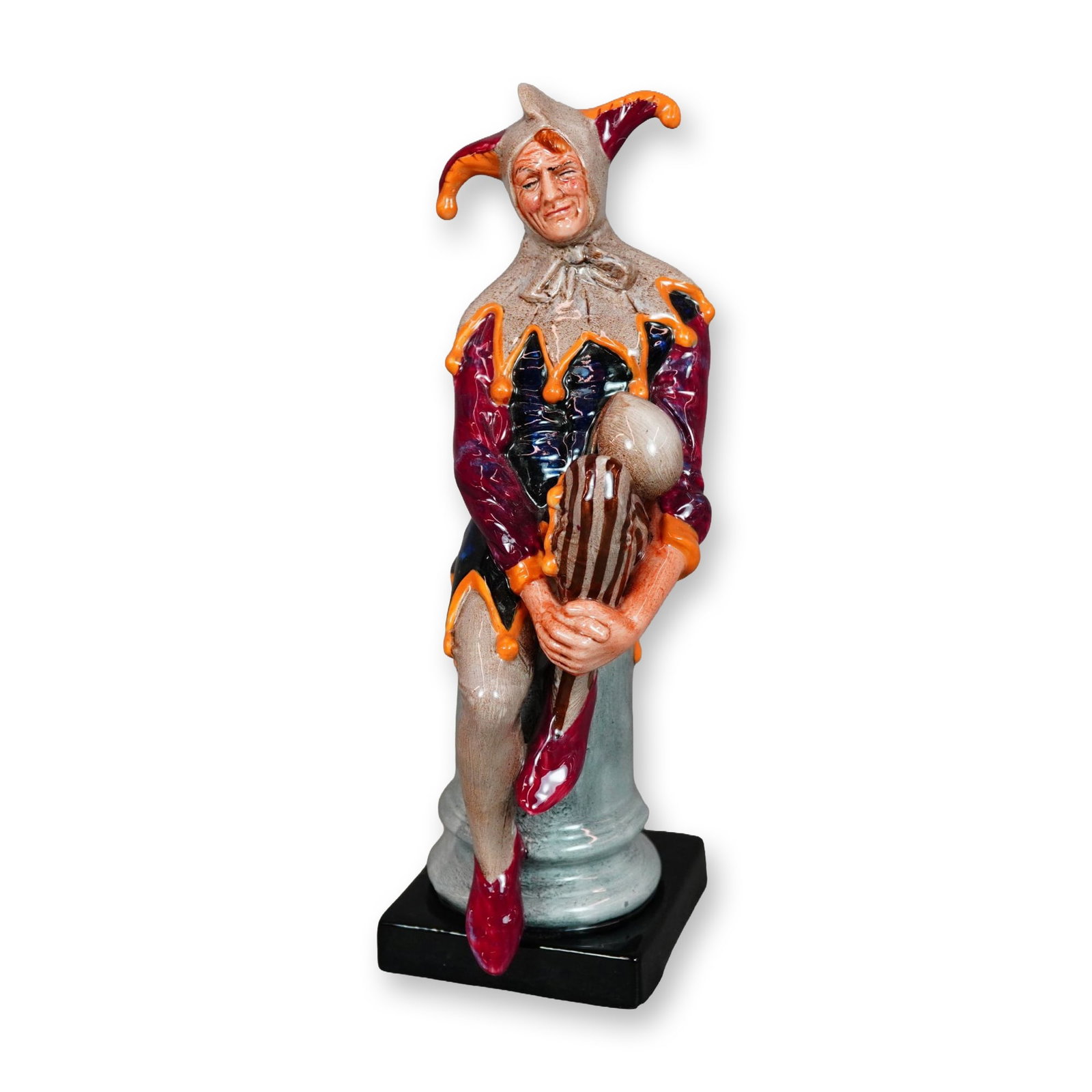 Royal Doulton Porcelain Figurine The Jester HN 2016: Royal Doulton porcelain figurine titled The Jester, model HN 2016. The figure depicts a seated court jester perched on a pale blue-gray classical column set on a square black plinth. The jester is dre