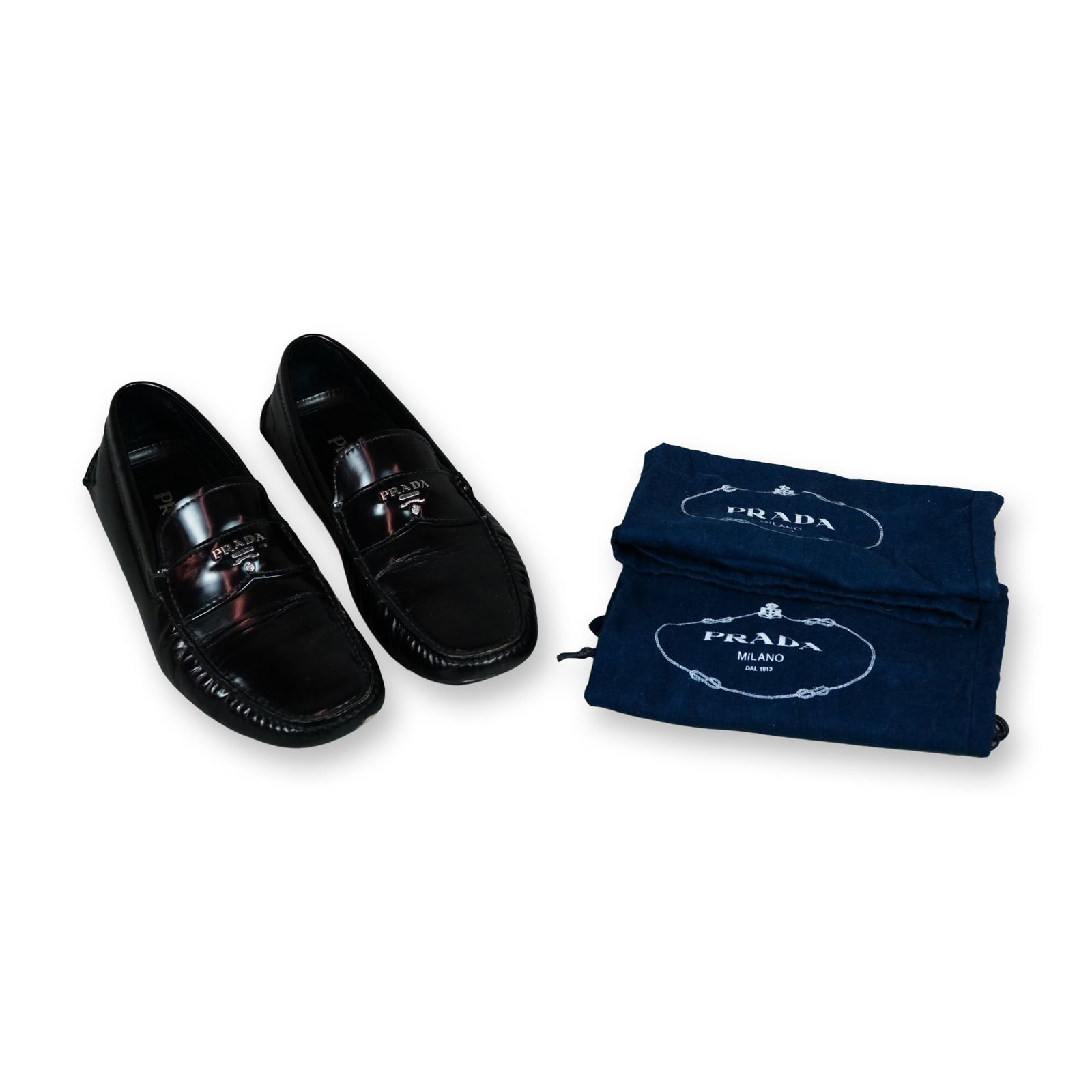 Prada Black Leather Driving Loafers: A pair of Prada model 2D2170 driving loafers in black calfskin leather, featuring metal Prada logo lettering on the vamp straps. Designed as a classic Italian slip-on slipper shoe, the interior featur